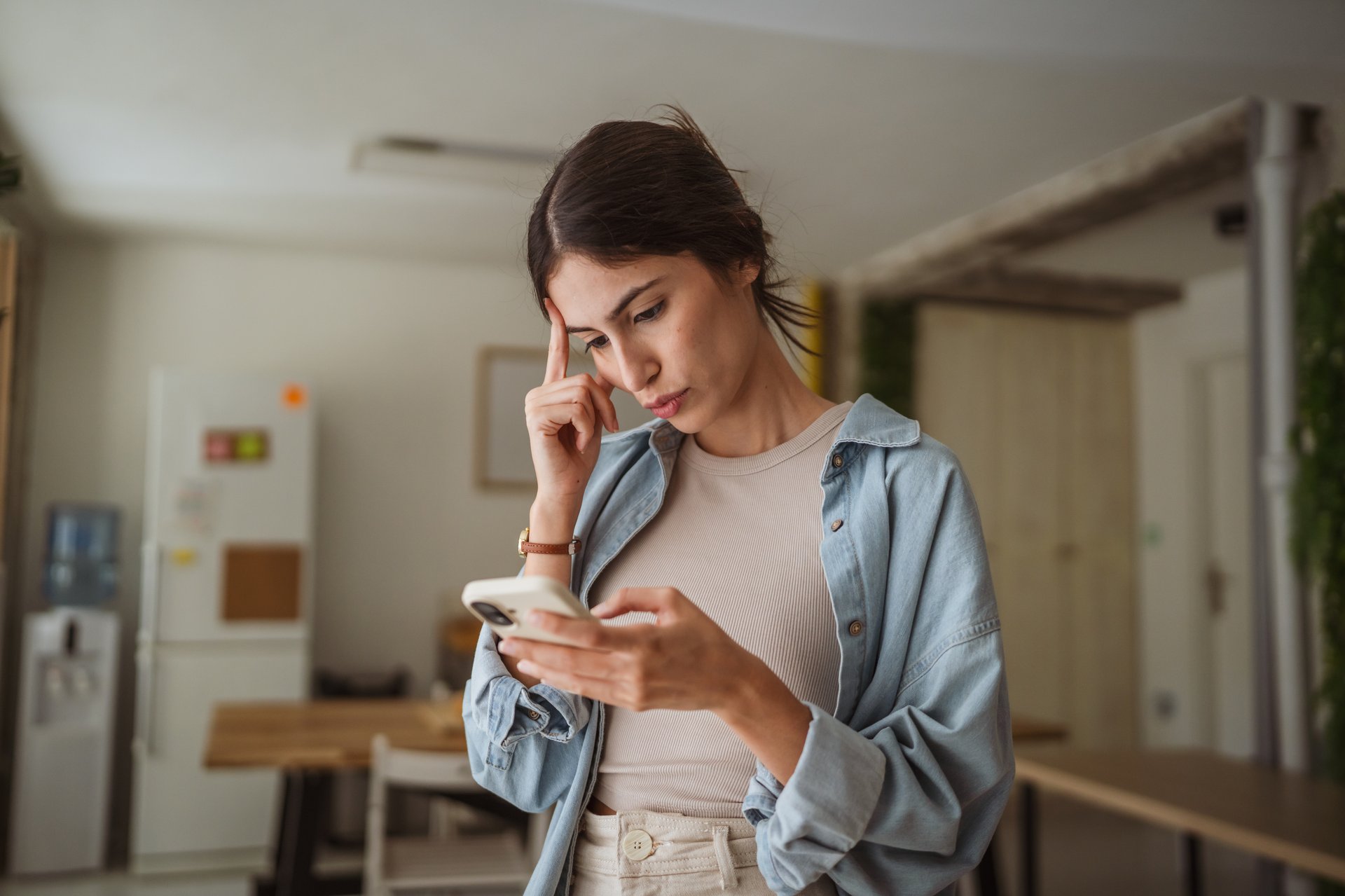 Young businesswoman is having problems while working from home and reading a message on her smart phone, touching her forehead with a thoughtful expression