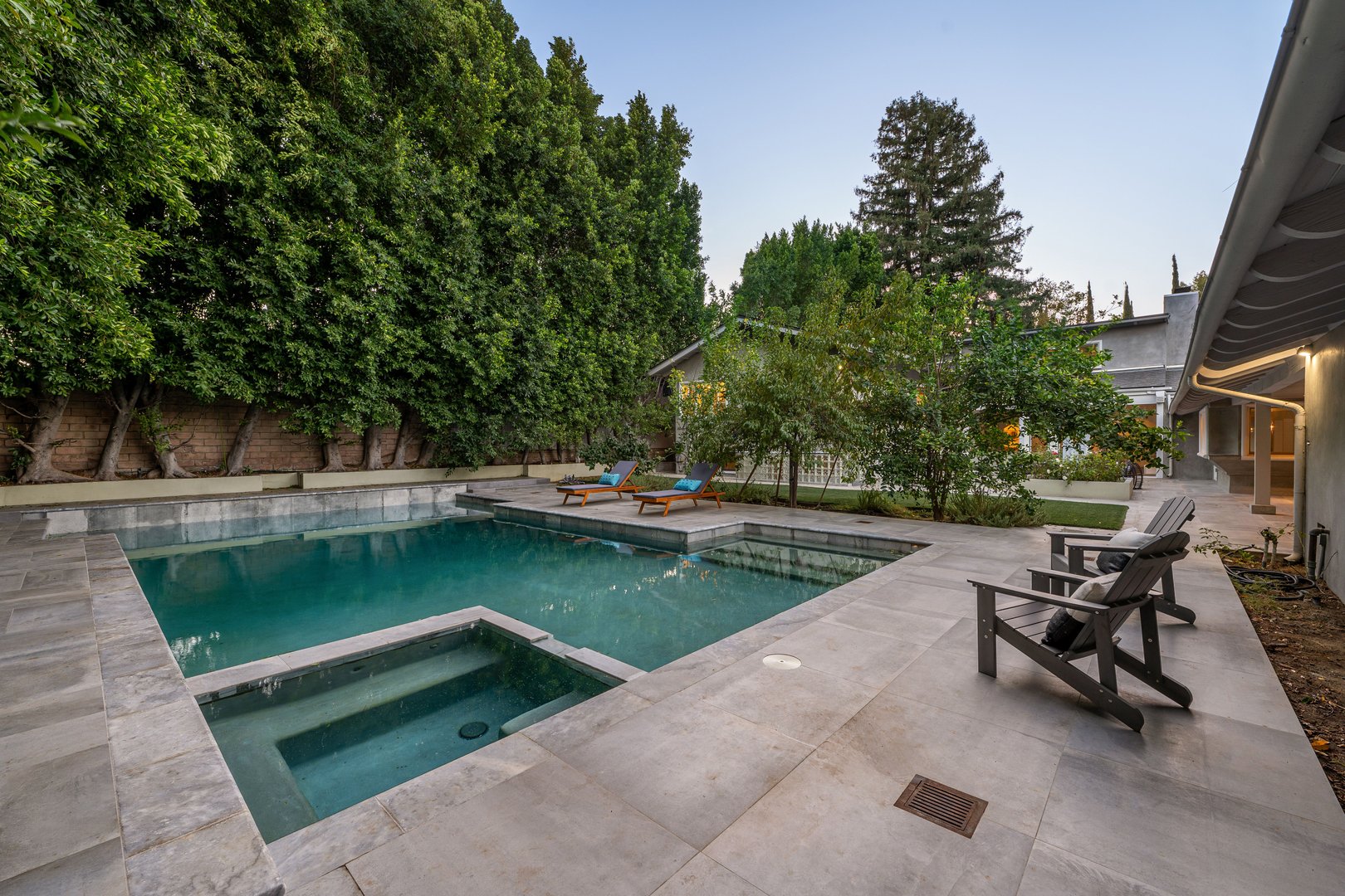 A modern backyard with a swimming pool, lounge chairs, and lush green trees