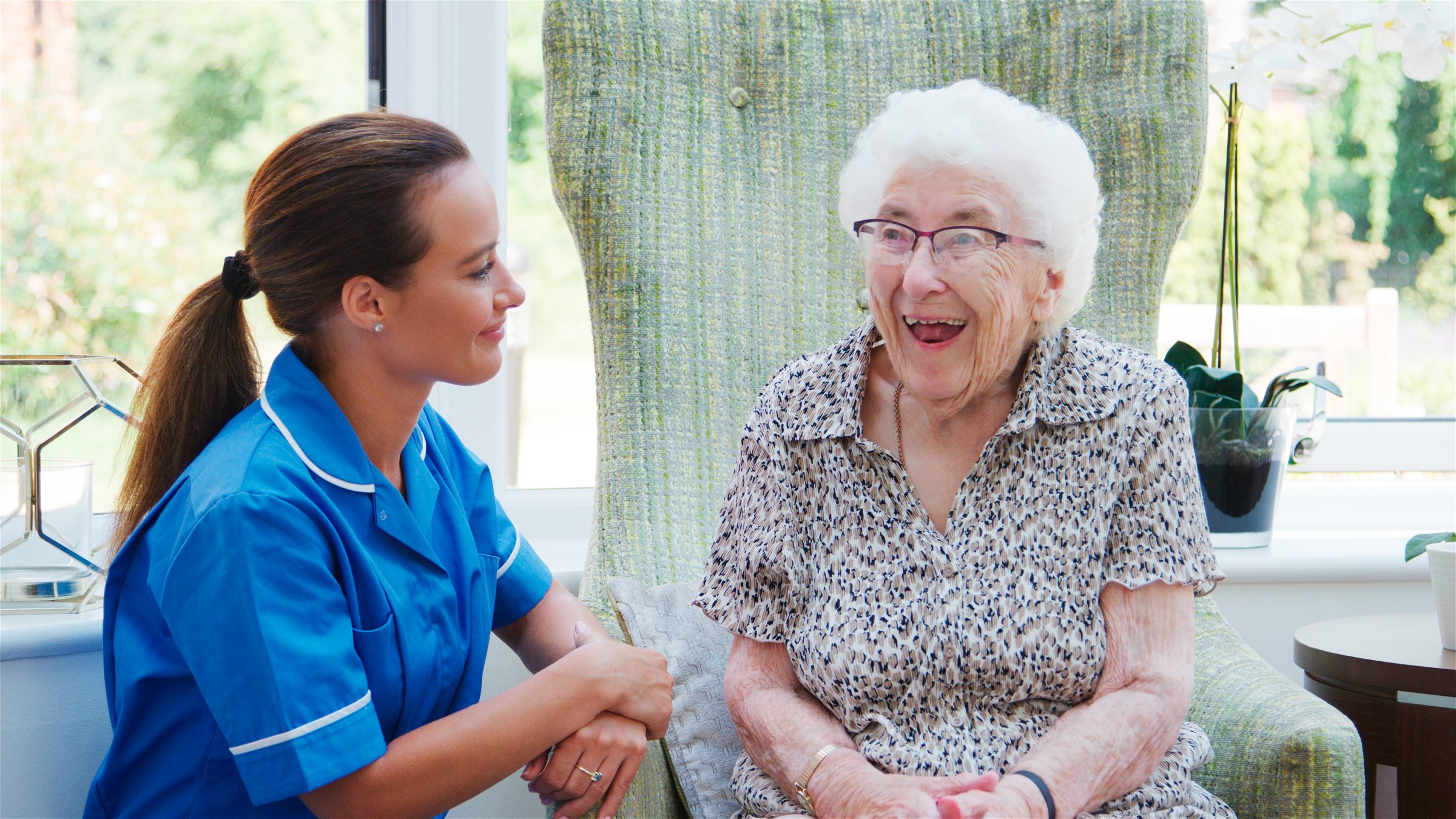 Laughing Senior Woman Sitting In Chair In Residents Lounge And Talking With Nurse In Retirement Home