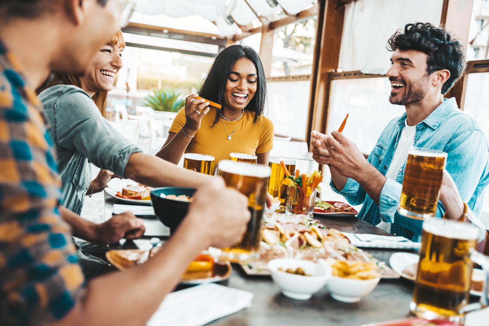 Multiethnic happy friends drinking beer glasses sitting at brewery pub restaurant table - Smiling guys and girls having fun at rooftop dinner party - Food and beverage lifestyle