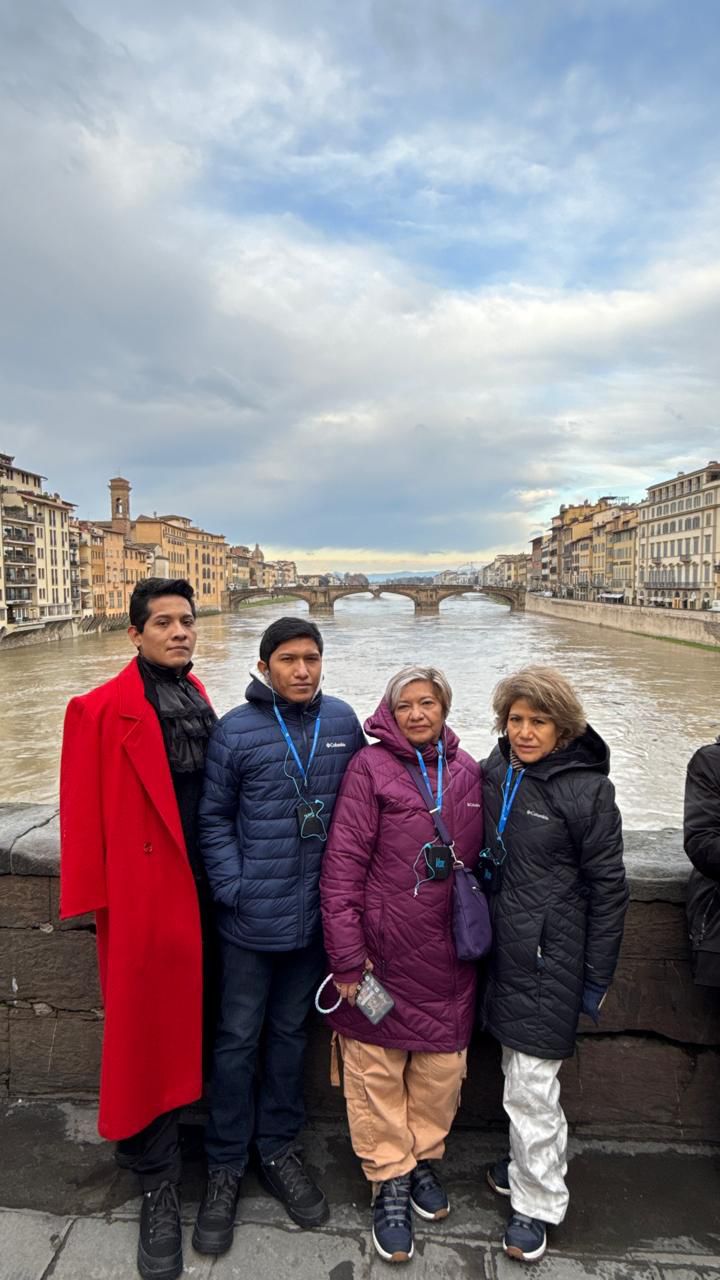 Four people bundled up in winter clothing stand on a bridge with a river and city buildings in the background on a cloudy day.