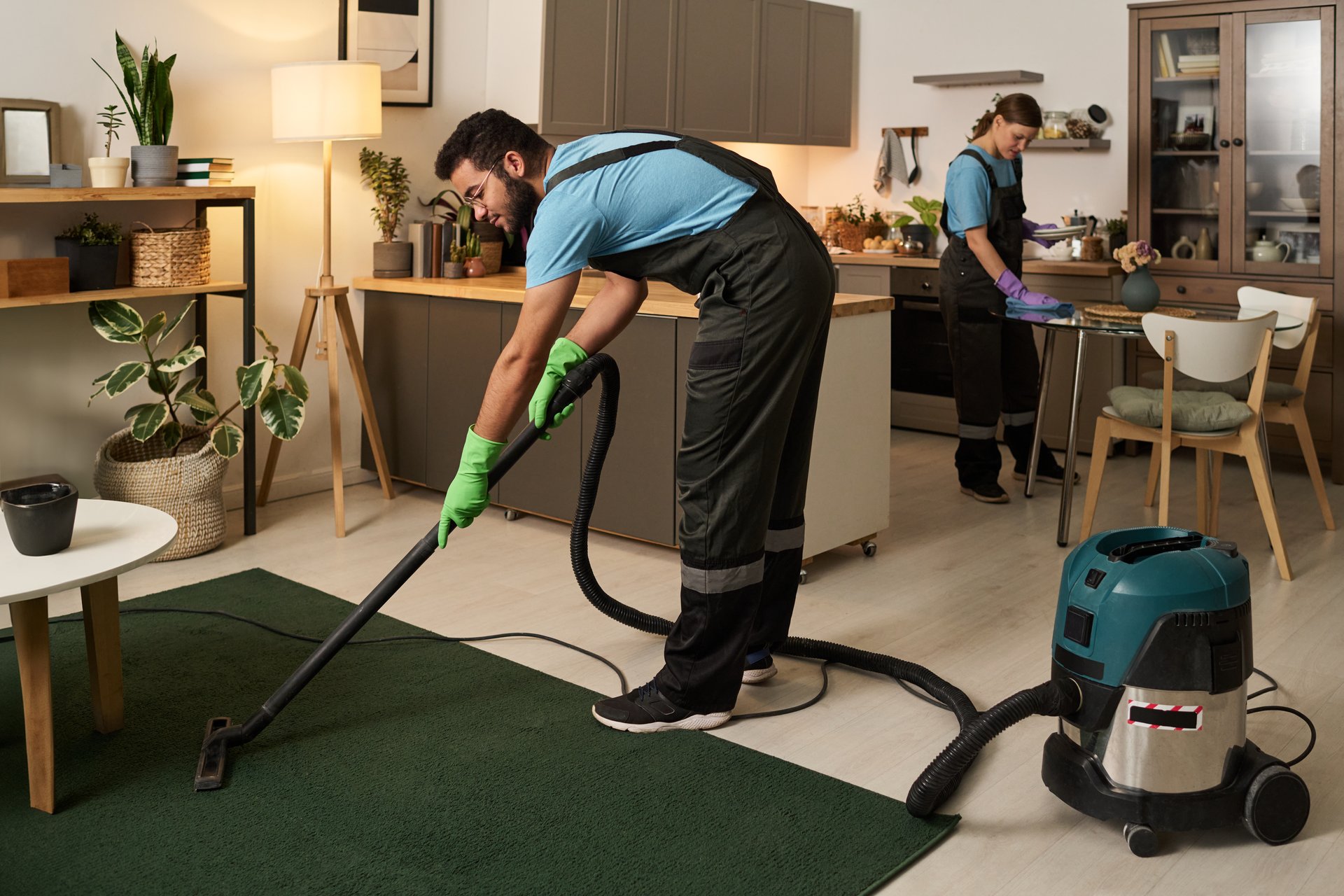 Man in uniform vacuuming carpet with vacuum cleaner together with his colleague wiping dust in background