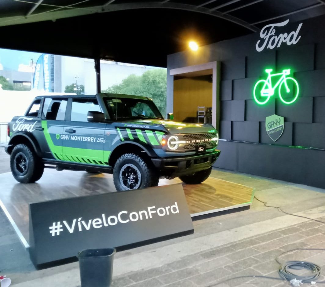 A Ford Bronco on display with green and black graphics, under a shelter, alongside a neon bicycle sign and #VíveloConFord sign.