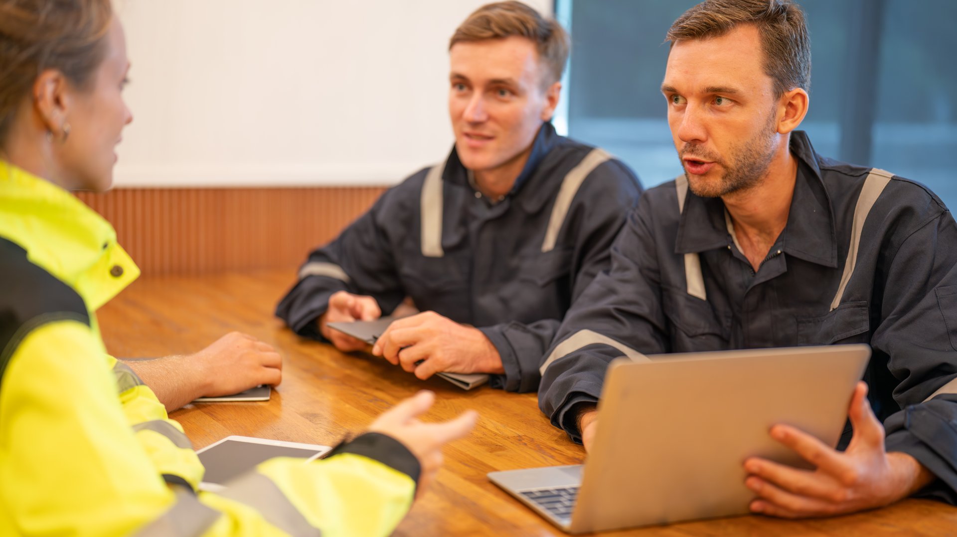 team of oil refinery engineers. wearing in safety wear, one man in casual wear, and another man in workwear discuss something at a table with a laptop. Business meeting, teamwork, and communication concept.
