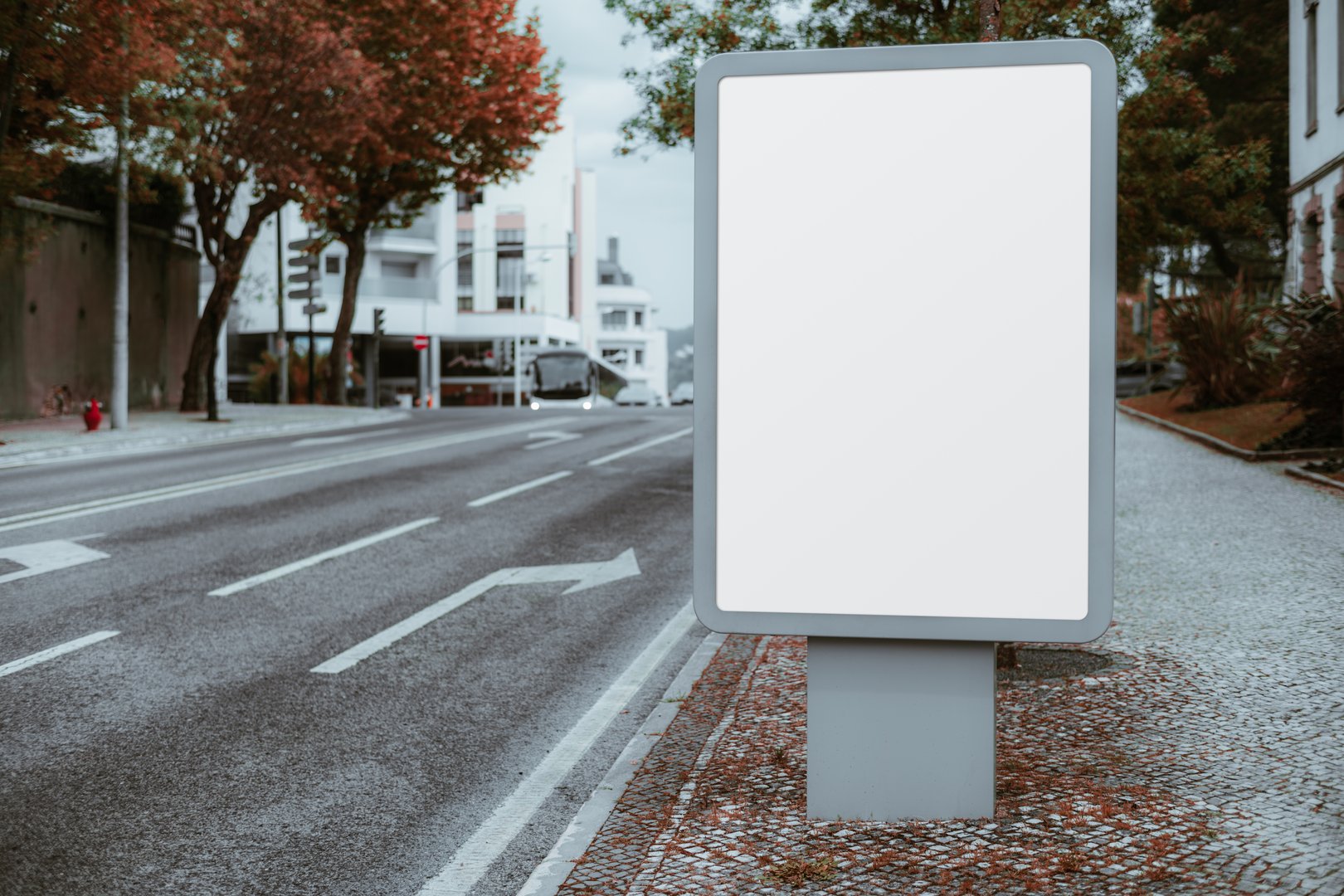An empty vertical billboard with a blank white screen stands on a cobblestone sidewalk next to a quiet urban road with no traffic, surrounded by trees and modern buildings
