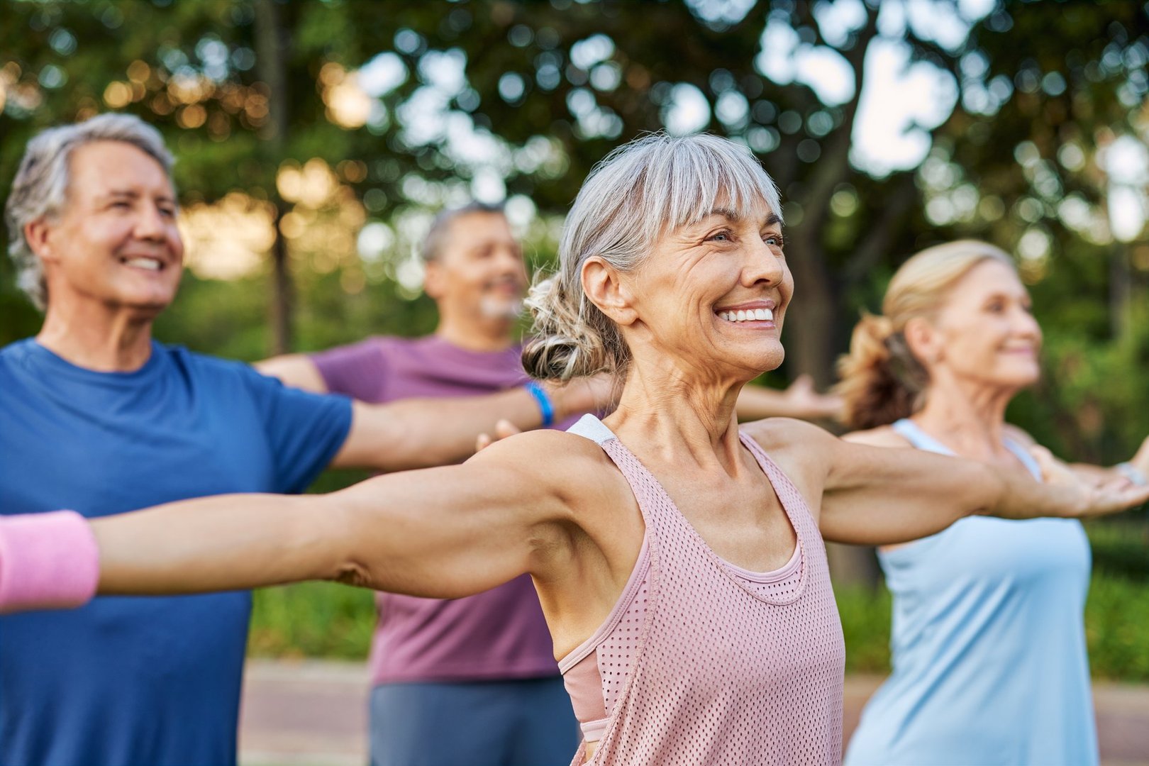 Happy senior group doing yoga together in the park, gentle gymnastics. Mature men and women stretching arms in outdoor fitness class. Sporty elderly friends doing a arm warm up workout before training class together in a garden.