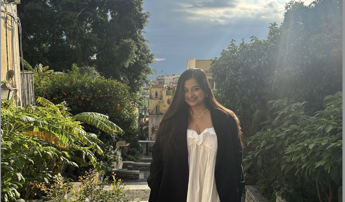 A woman in a white dress and black coat stands in a lush garden with sunlight streaming through the clouds.