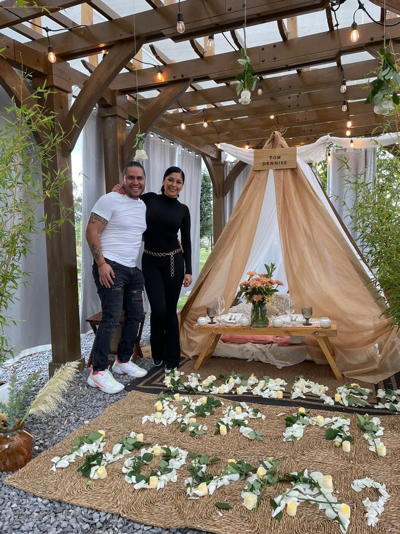 Couple posing beside a romantic tent with flowers and candles, under a wooden pergola with string lights.