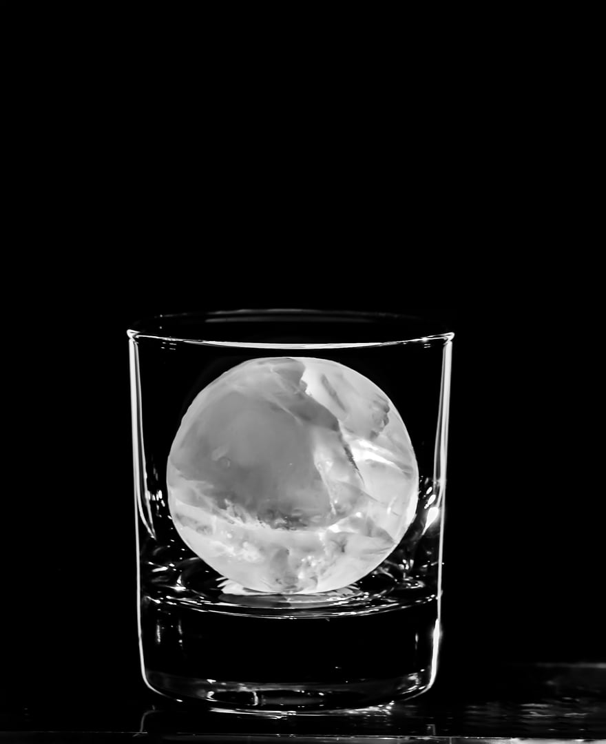 Round spherical ice cube ball in a cocktail glass with unique rim lighting against a black background.