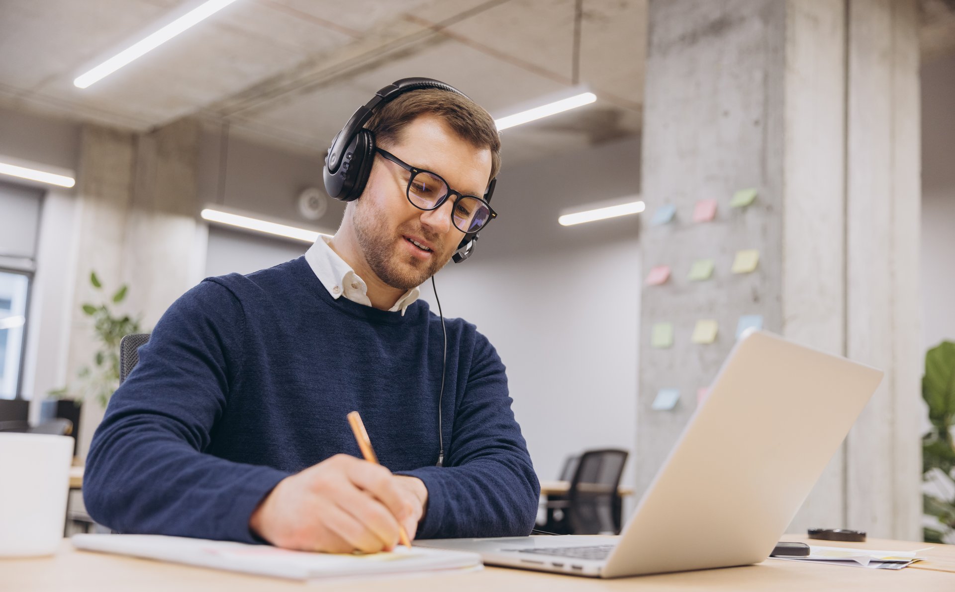 Office worker engaging in an online meeting while taking notes and using a laptop, showcasing a modern workspace filled with technology