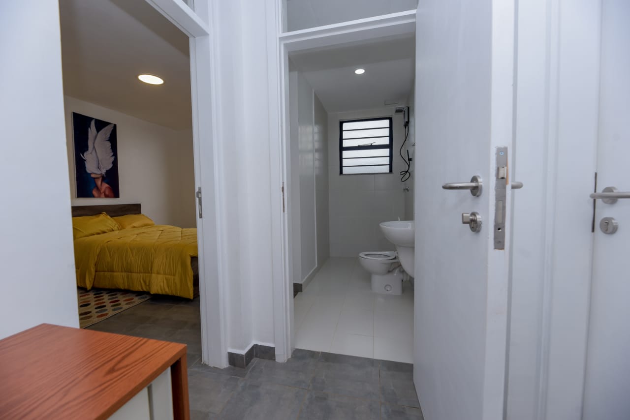 A hallway view of an apartment with a bathroom and bedroom. The bedroom has a yellow bedspread and a framed artwork on the wall.