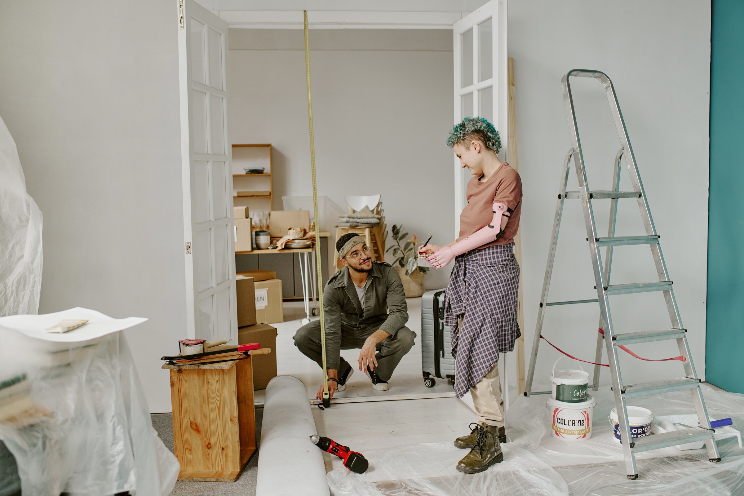 Young adult man kneeling on floor measuring distance while young adult woman standing nearby holding notepad discussing renovation in new home interior
