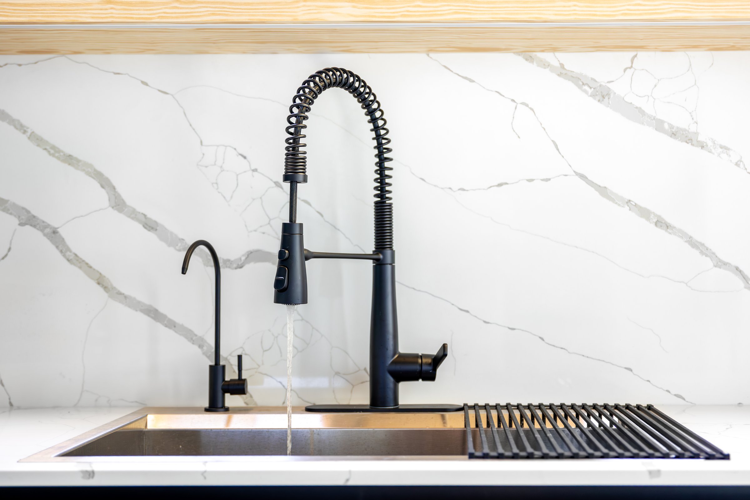 Water flows from a black professional faucet over a steel sink against a white marble wall. Modern kitchen furniture with crisp geometry and a premium feel.