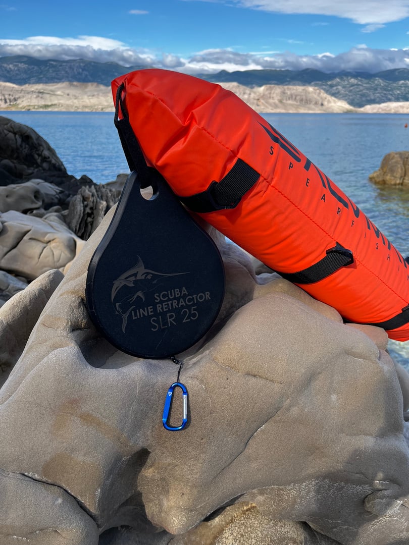 Scuba line retractor and bright orange float on rocky shore with ocean and mountains in background.