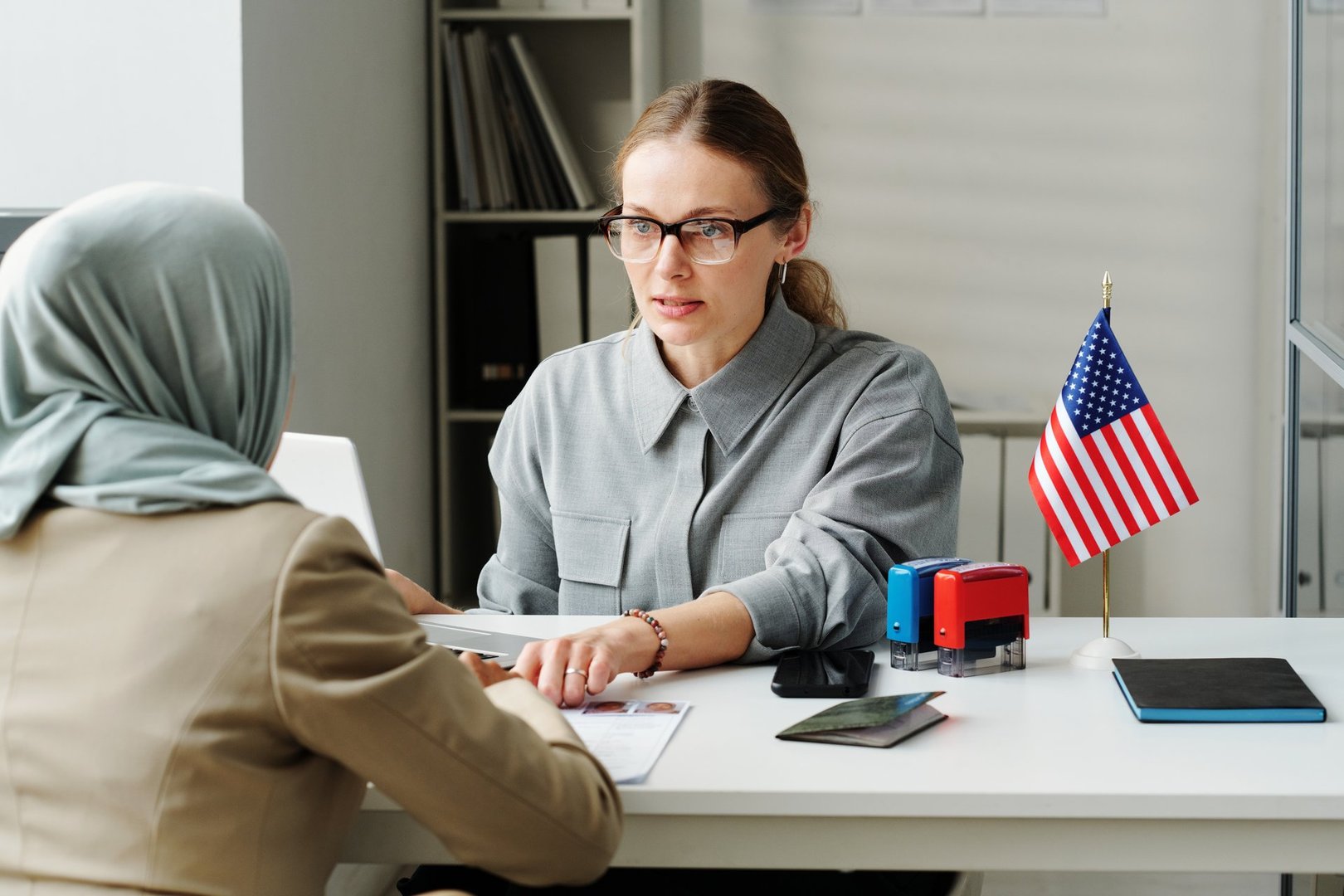 Modern Caucasian consular officer sitting at desk in front of Muslim woman explaining her how to fill in application form