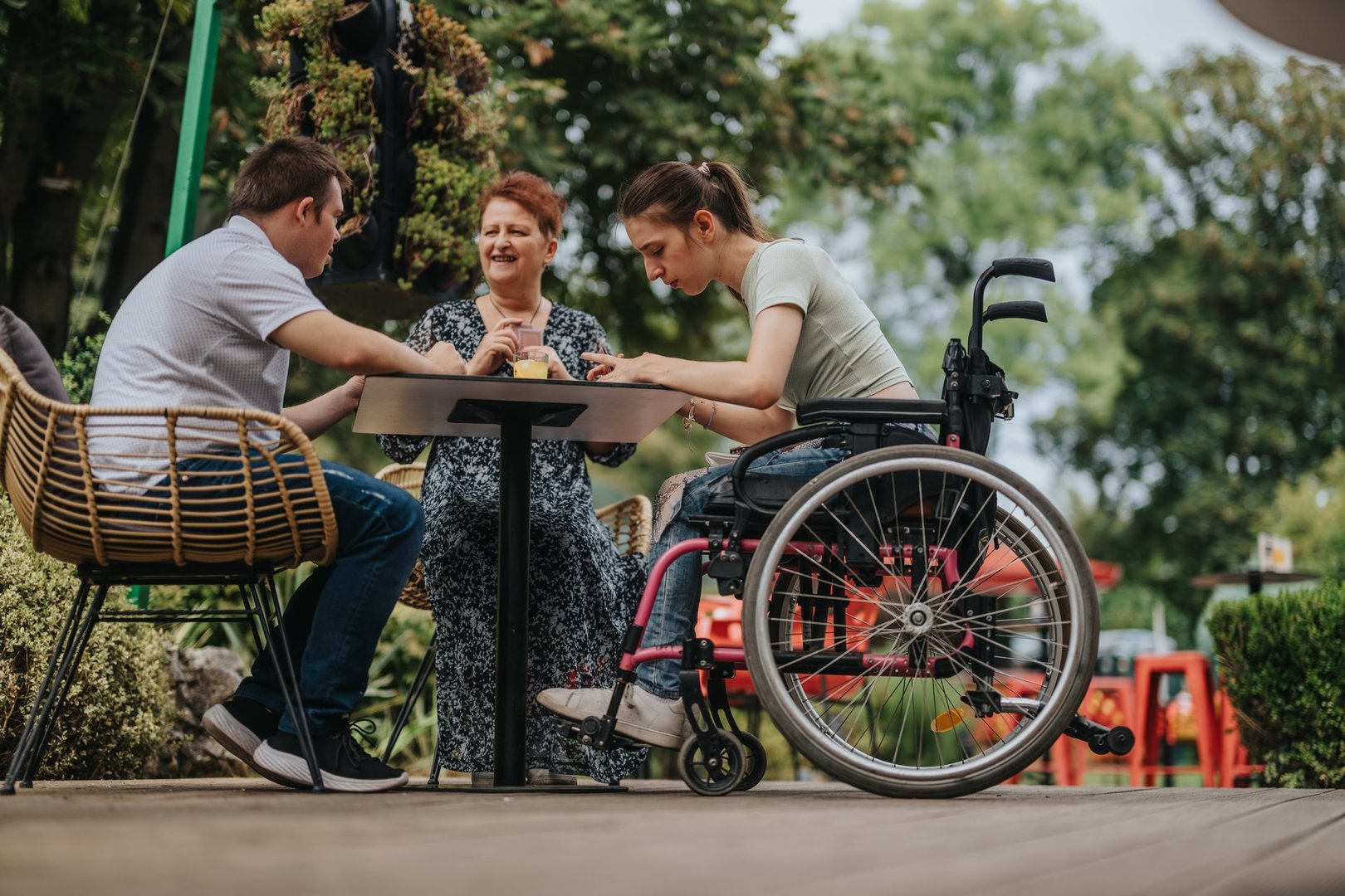 Three friends, one of whom uses a wheelchair, share a meaningful moment at an outdoor table in a lush green environment, emphasizing the strength of friendship and the importance of inclusivity.
