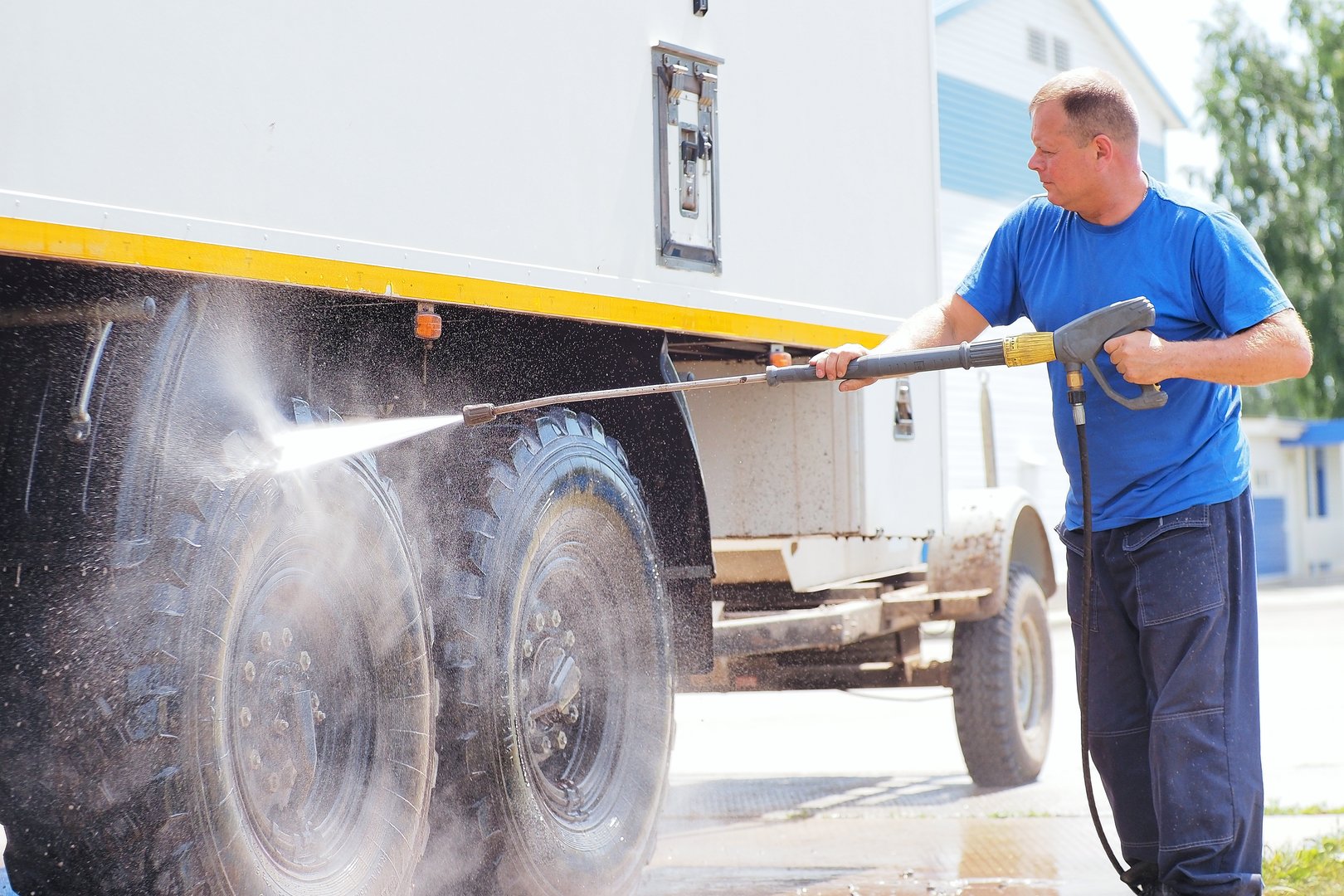 Man in overalls washes truck with water gun. Car wash for trucks. Car washer sprays car with pressurized water jet on summer day.