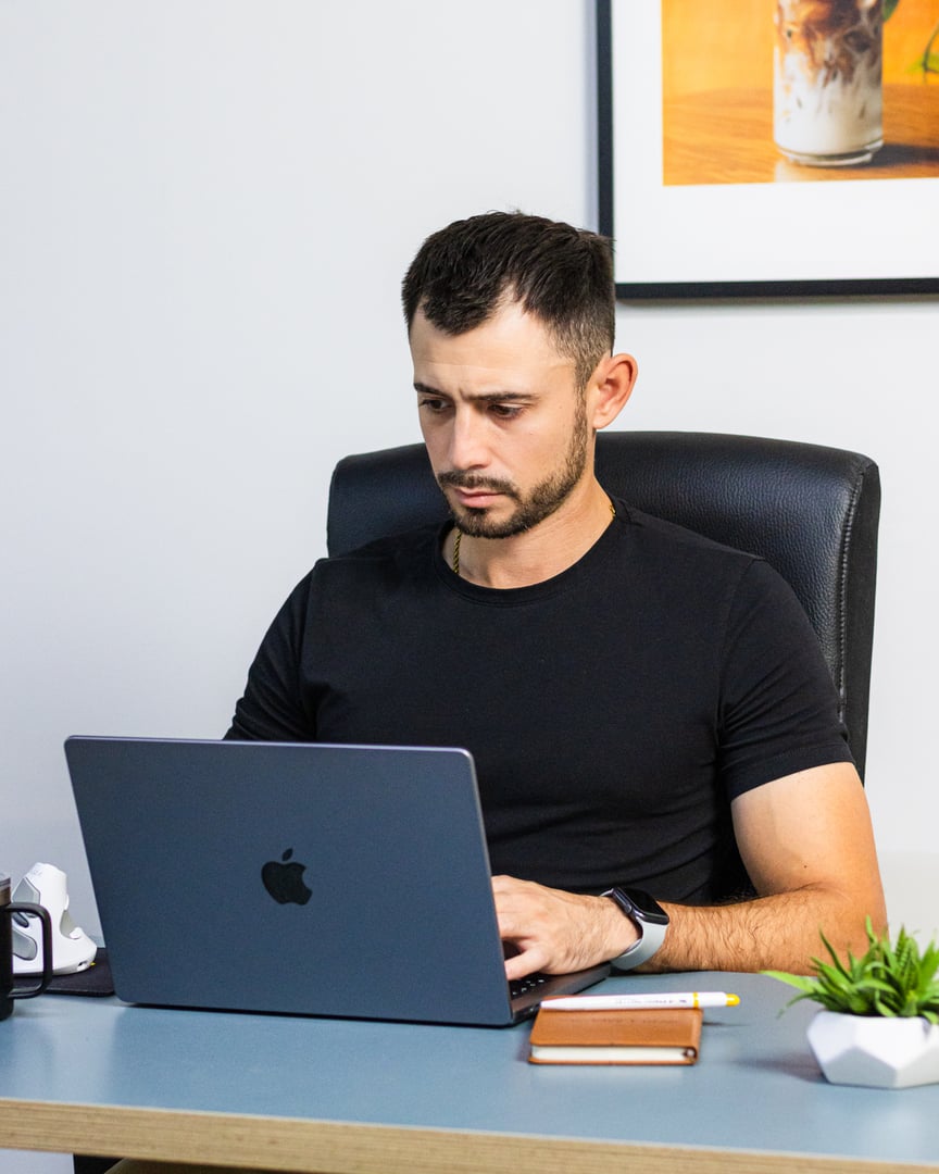 Man in a black shirt working on a laptop at a desk with a plant and notebook, seated in an office environment.