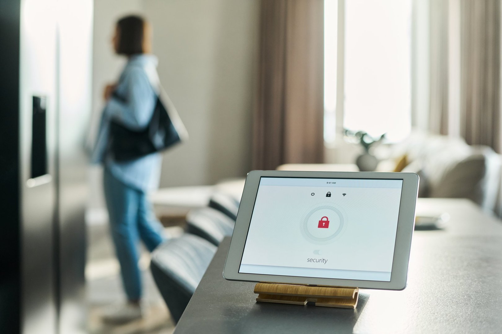 Tablet displaying security system interface on kitchen counter with blurred Caucasian young adult woman standing in background holding bag and looking away from camera