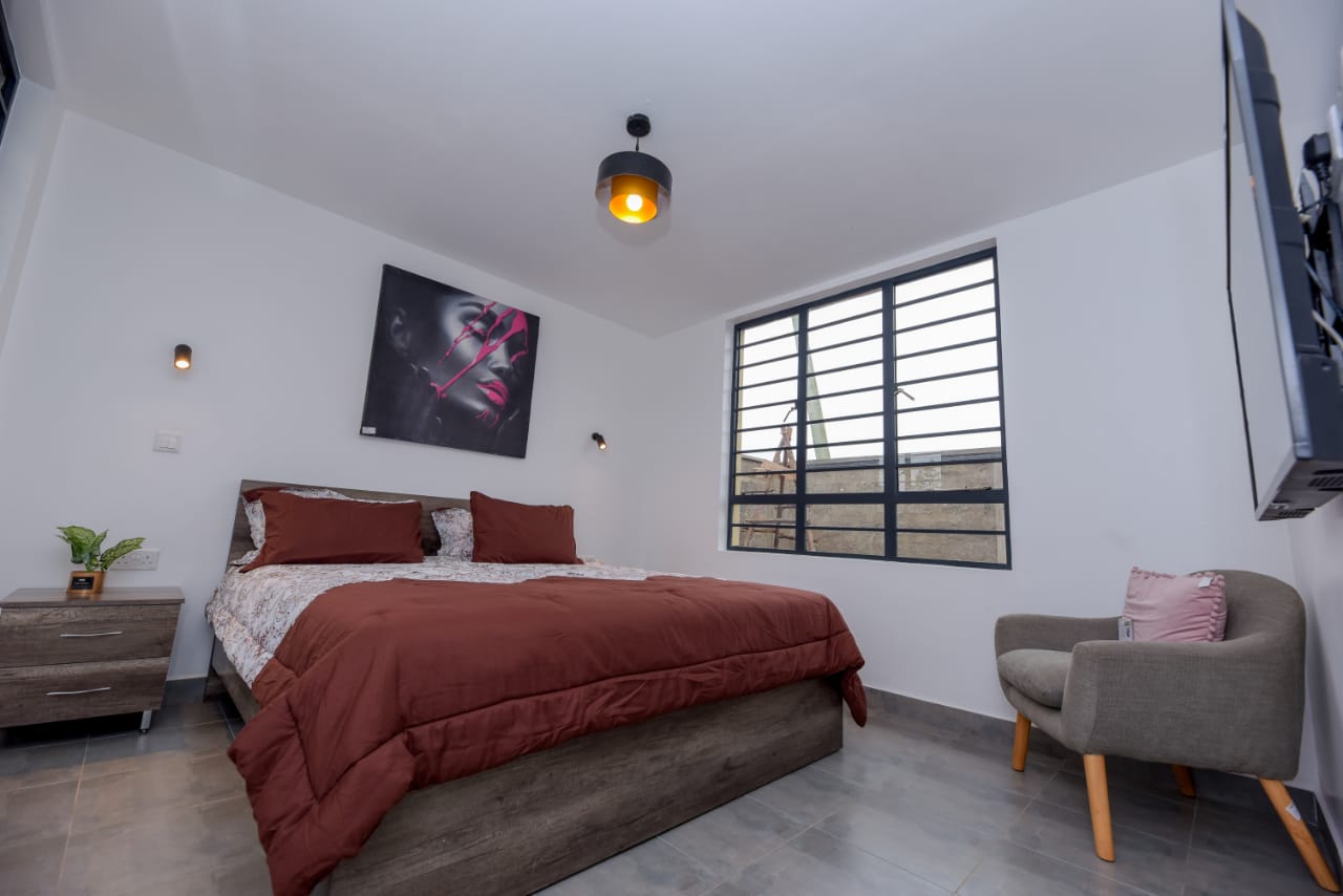 Minimalist bedroom with a double bed, brown bedding, wall art, a side table, and a cushioned chair by the window.
