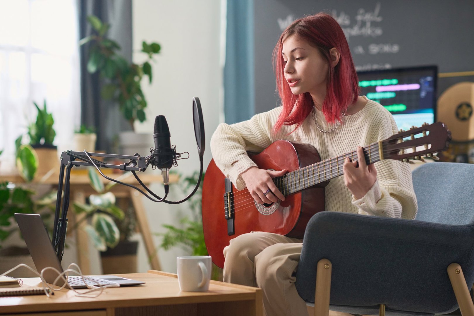 Pretty girl in casualwear performing new song and playing acoustic guitar while sitting in front of laptop and singing in microphone