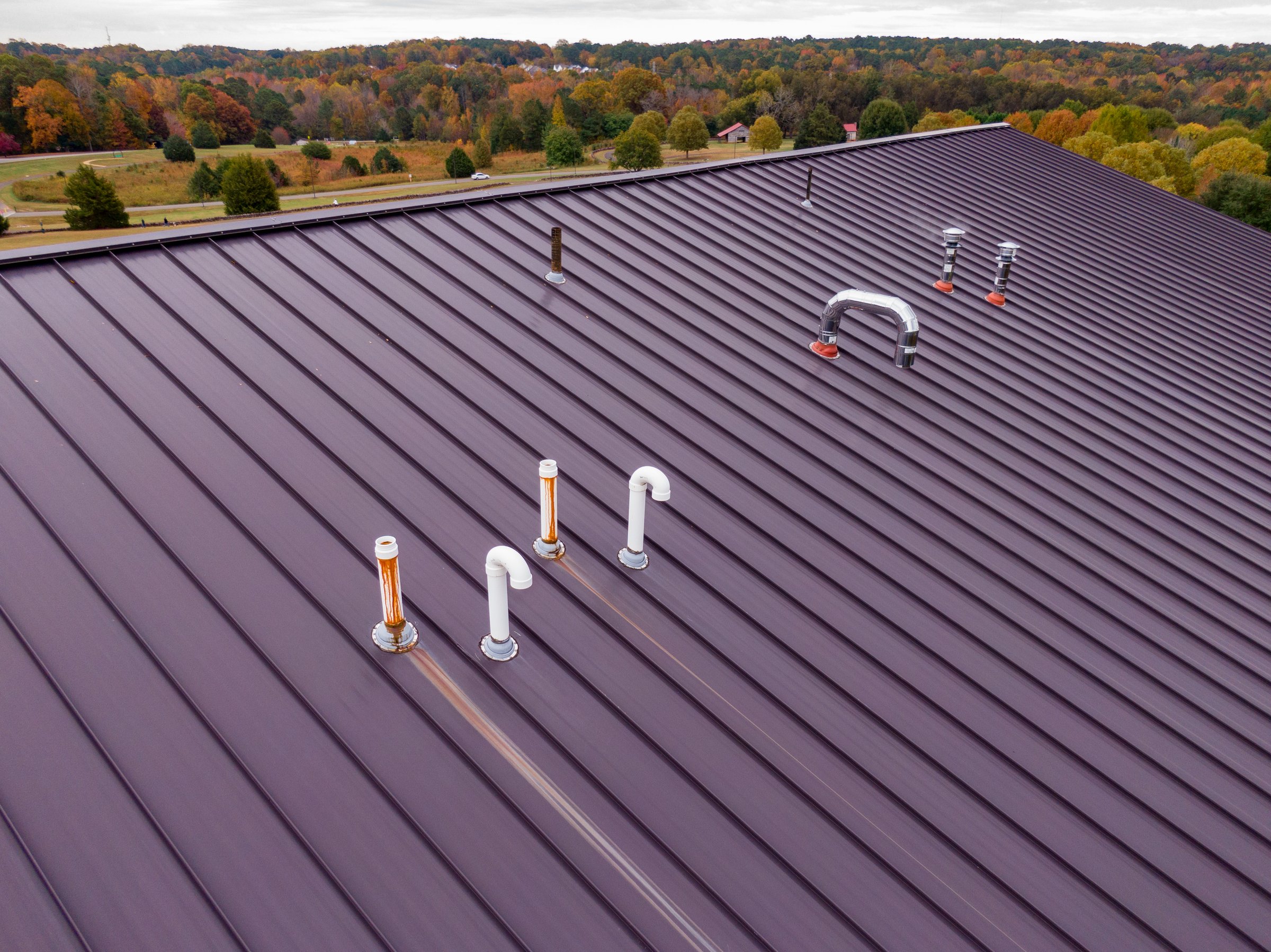 A high quality aerial close-up captured by a drone, detailing a dark brown or bronze-colored standing seam metal roof on a modern commercial or institutional building. The image highlights the clean, durable lines of the roofing material, including various vents, flashing, and mechanical components. In the background, the beautiful fall foliage of the surrounding park (Joyner Park, Wake Forest, NC) provides a natural contrast. This image is ideal for use in construction, architecture, and building material catalogs.