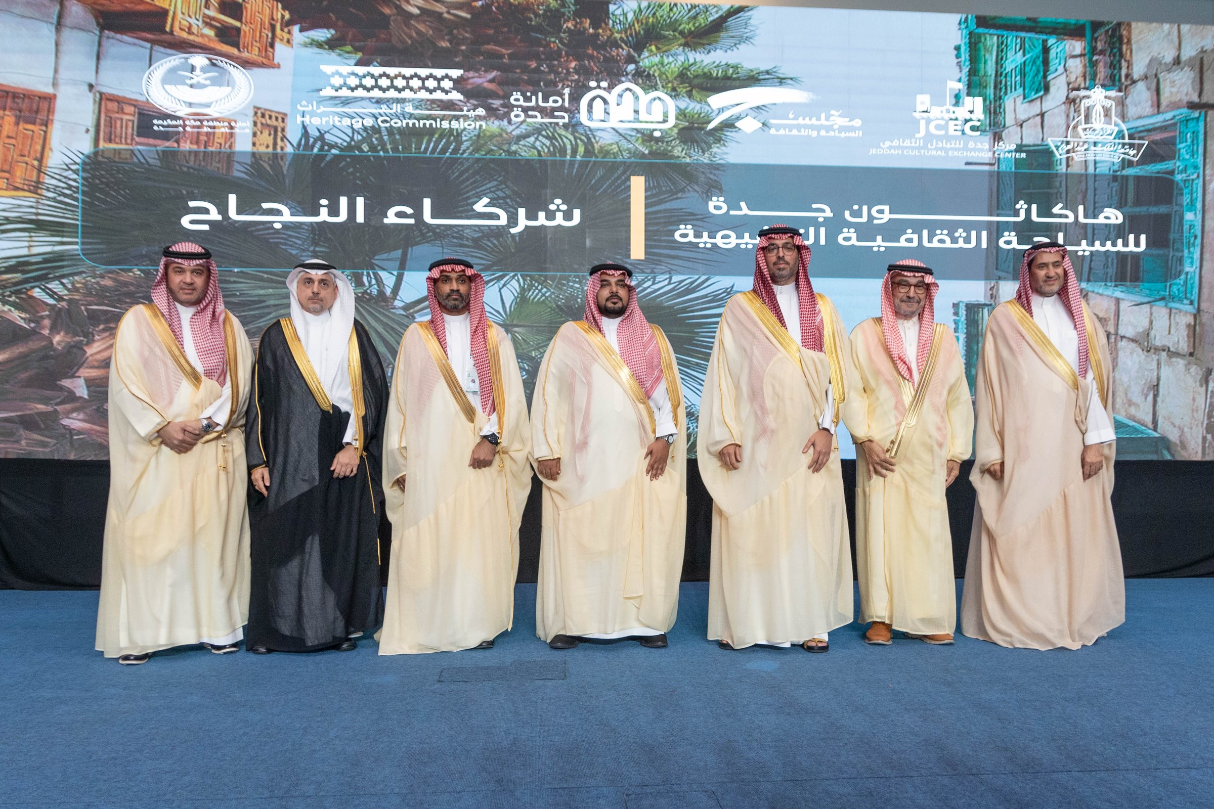 A group of Arab men in traditional attire stand on stage with a backdrop featuring logos and Arabic text.