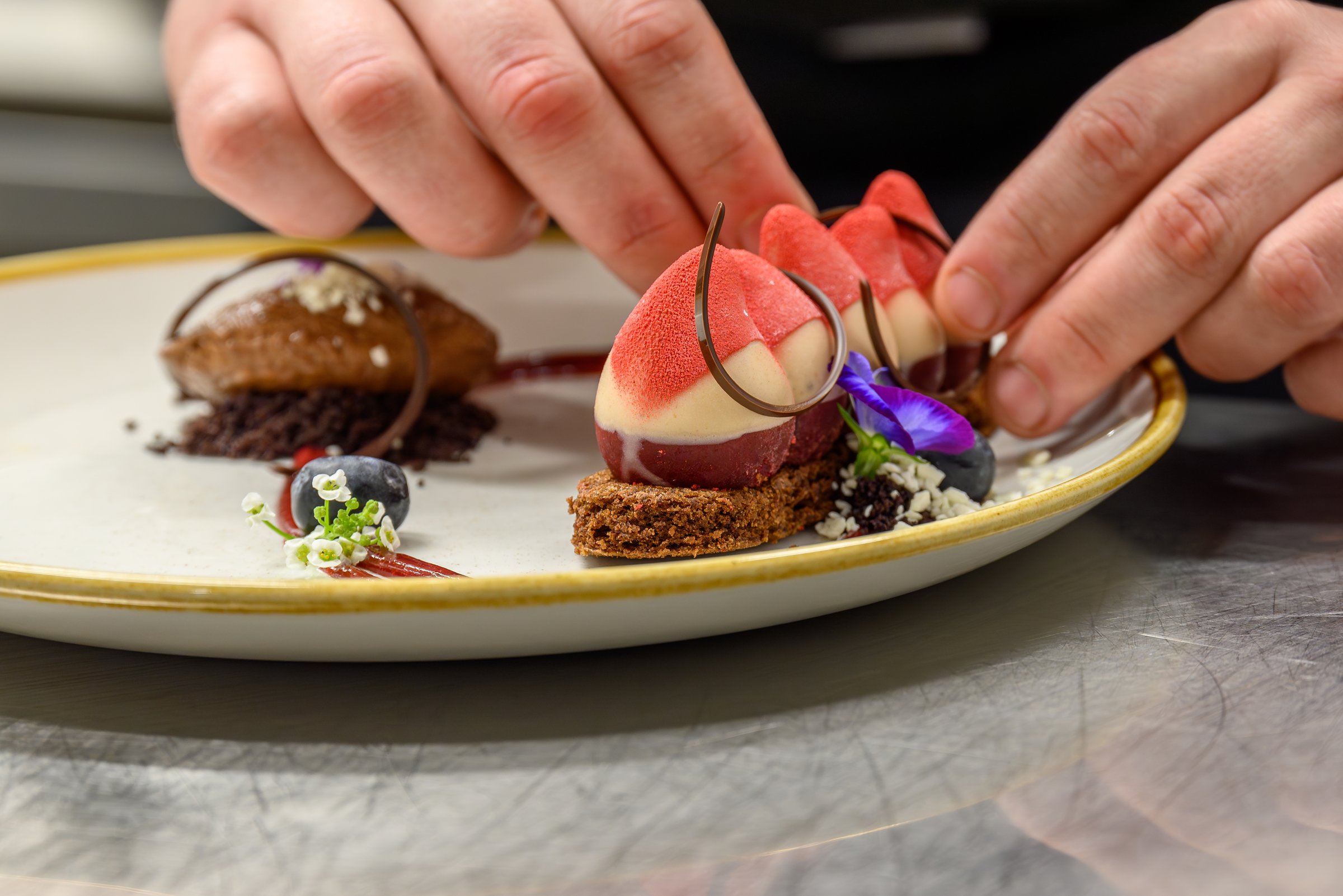 A close-up of an elegant dessert being artfully plated by a chef. The dish features mousse, delicate chocolate decorations, vibrant sauces, and edible flowers, showcasing the creativity of fine dining desserts.