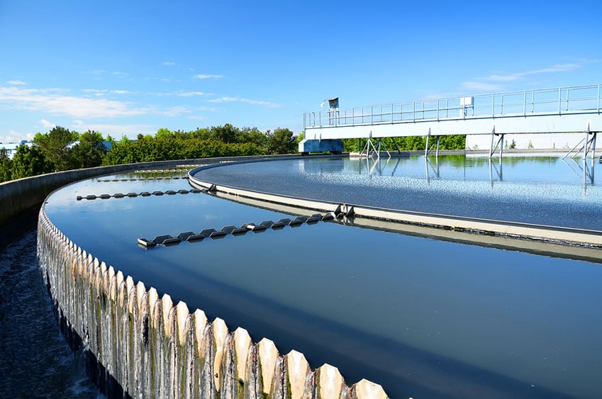 Circular wastewater treatment tank with clear blue sky, trees in the background, and water flowing over the edge.