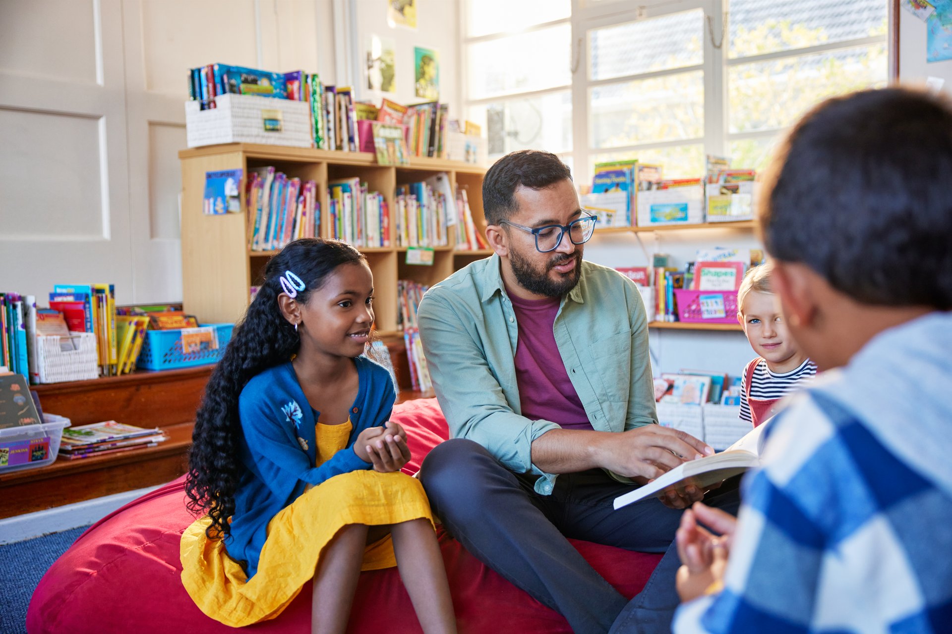 Male teacher reading a book to multiethnic students while sitting in school library in a circle on big soft pouf. Volunteer teacher reading a fairy tale to a group of happy elementary children. Librarian at primary school reading to children at story time and explains the moral of it.