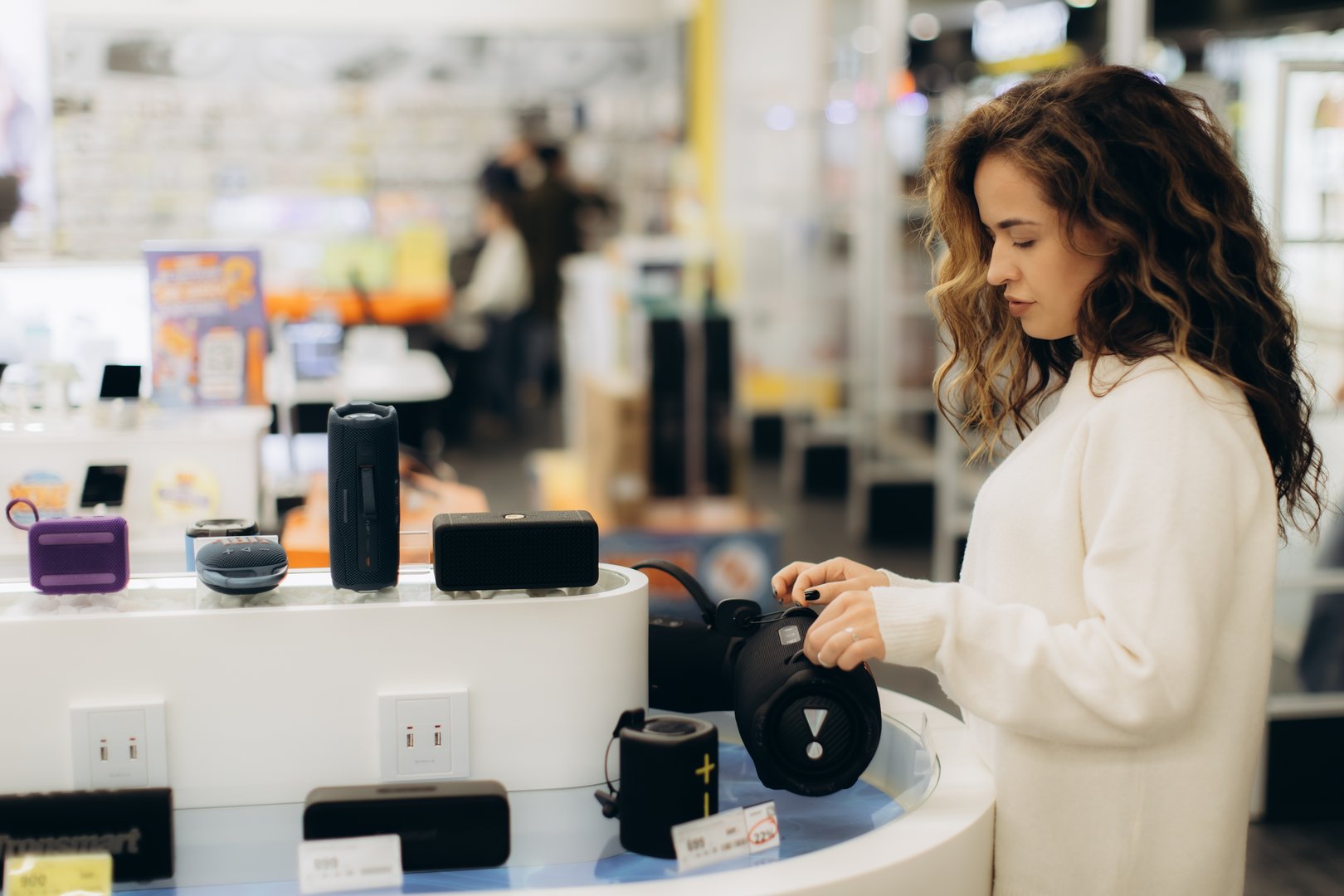 A young woman explores a variety of portable speakers in a well-lit electronics store, evaluating options for purchase amid an organized display of gadgets and equipment.