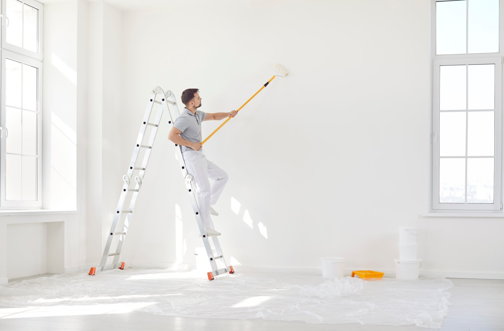 Male painter or construction worker painting wall surface in white color with paint roller brush, service man on stepladder finishing task for room, building repair, interior architecture and design