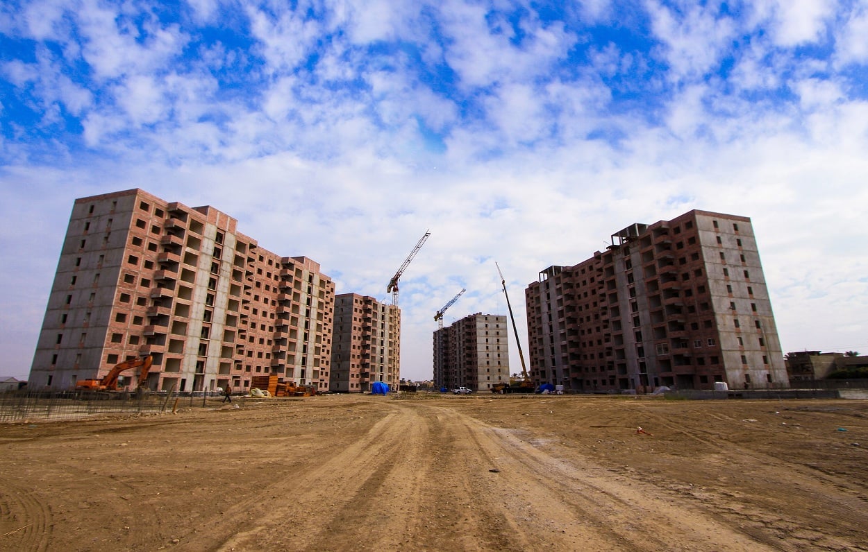 Two rows of unfinished apartment buildings with cranes and construction equipment, under a blue sky with clouds.