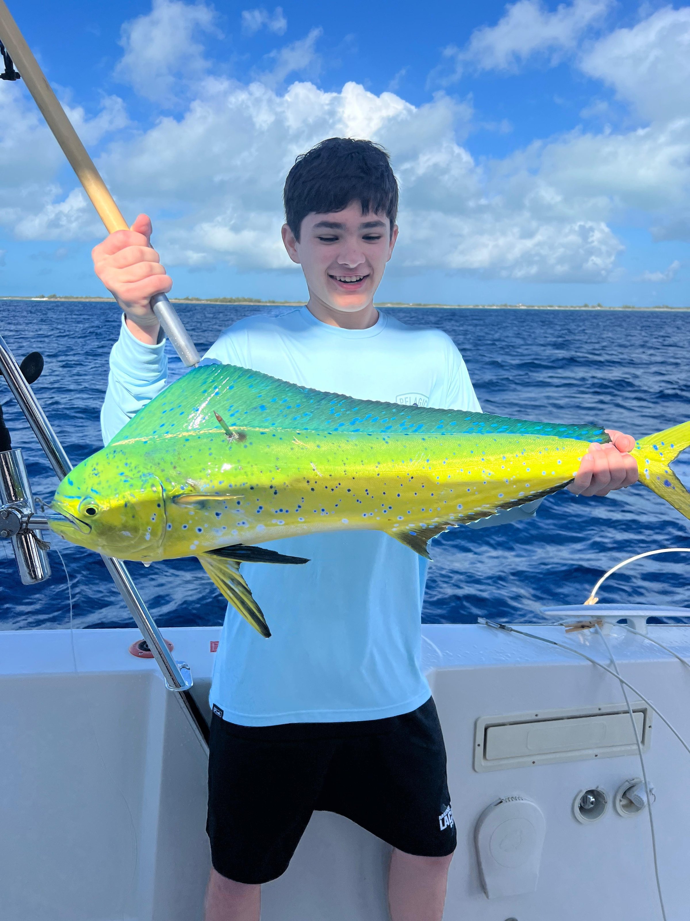 Teenage boy holding a large, colorful fish on a boat with a cloudy sky and ocean in the background.
