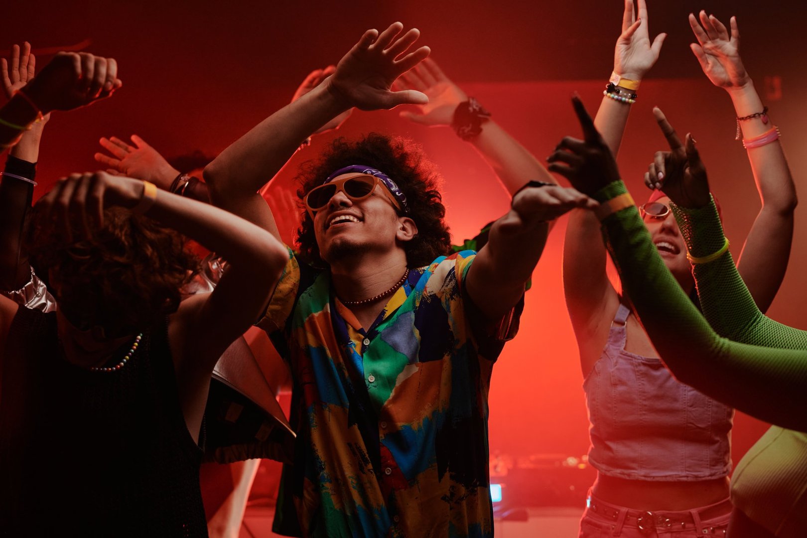 Young excited man in stylish apparel and trendy sunglasses keeping his arms raised while dancing at disco party among his friends