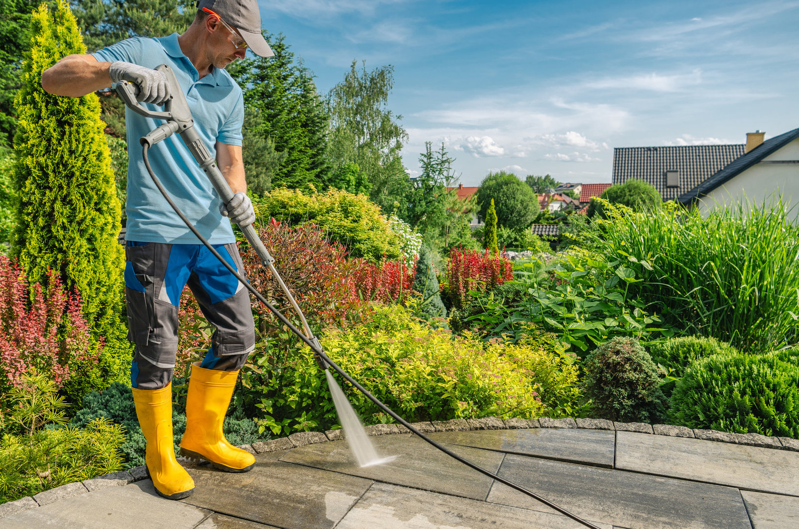 A man in yellow rubber boots operates a pressure washer to clean a patio surrounded by vibrant greenery and colorful flowers under a clear blue sky.