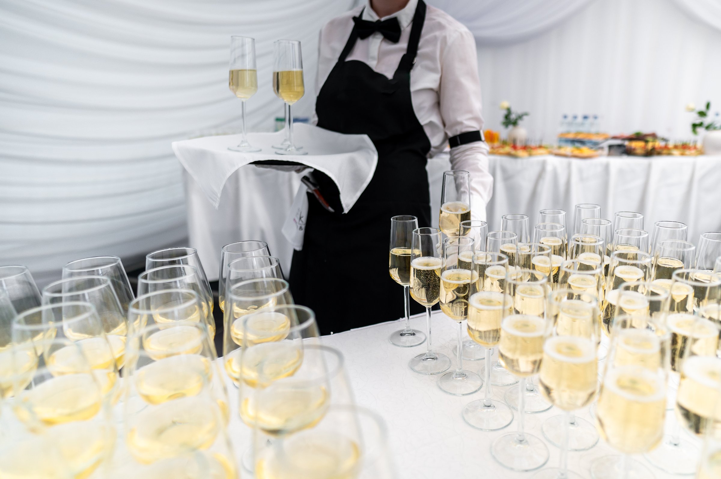 A waiter in a black vest and white shirt holds a tray of champagne glasses at an elegant event. In background, a table is set with various appetizers, creating a sophisticated and inviting atmosphere.