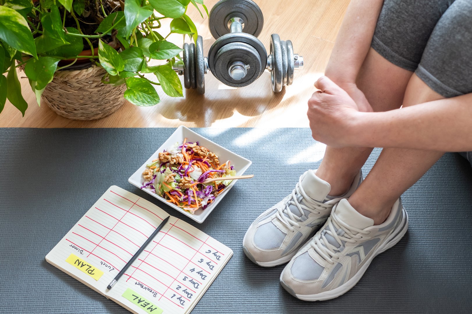 Mature woman sitting on a yoga mat at home, planning her healthy meal and workout routine, with a salad, dumbbells, and a meal plan notebook