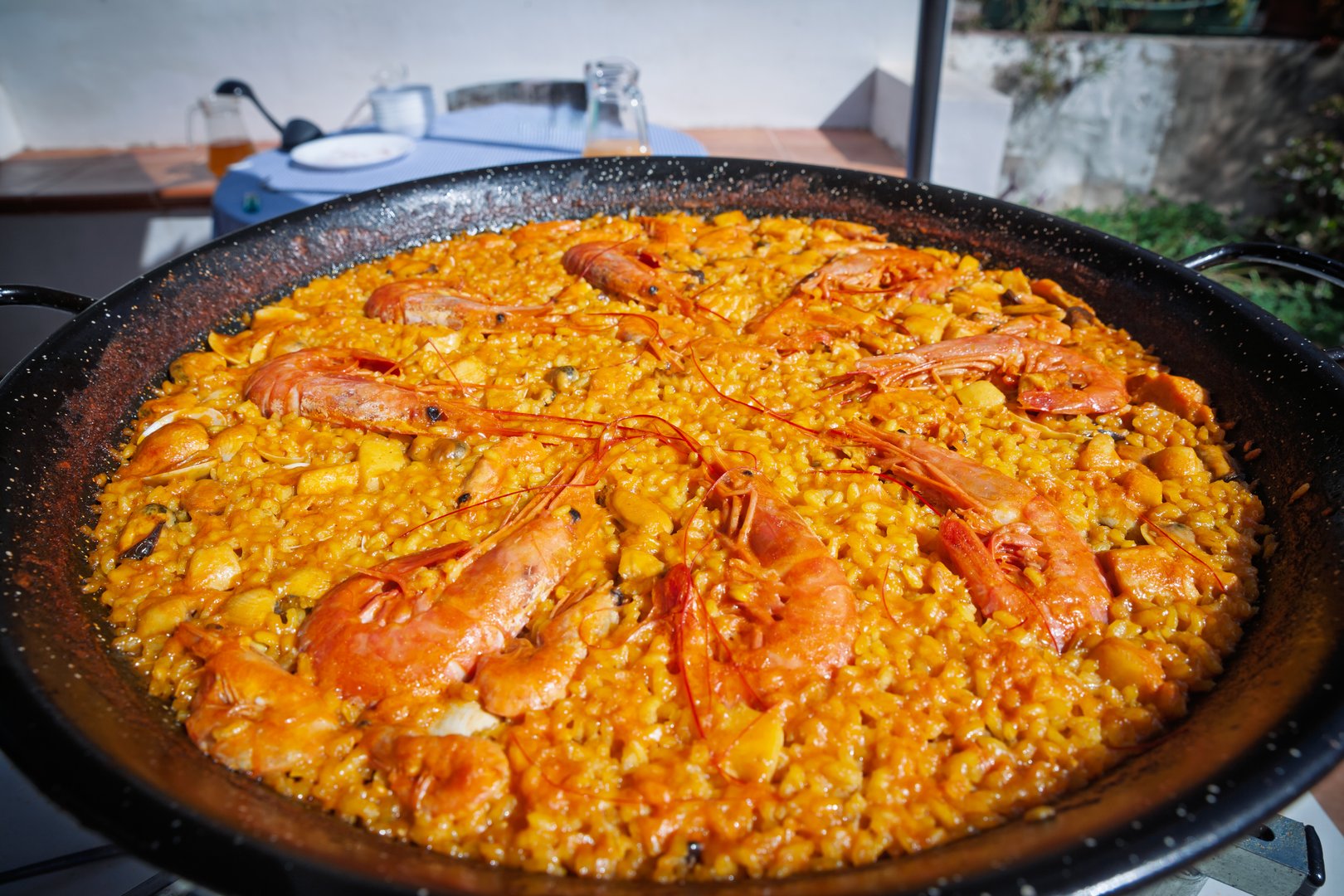 High-resolution close-up of a freshly cooked 'paella de mariscos'the classic Valencian seafood paellapresented in a wide black steel pan. Golden saffron rice is studded with mussels, squid and diced fish and crowned with whole red prawns arranged in a star pattern. The glistening surface hints at rich stock and extra-virgin olive oil. Soft background blur shows an outdoor table, suggesting home cooking or restaurant terrace dining on a sunny Mediterranean day.