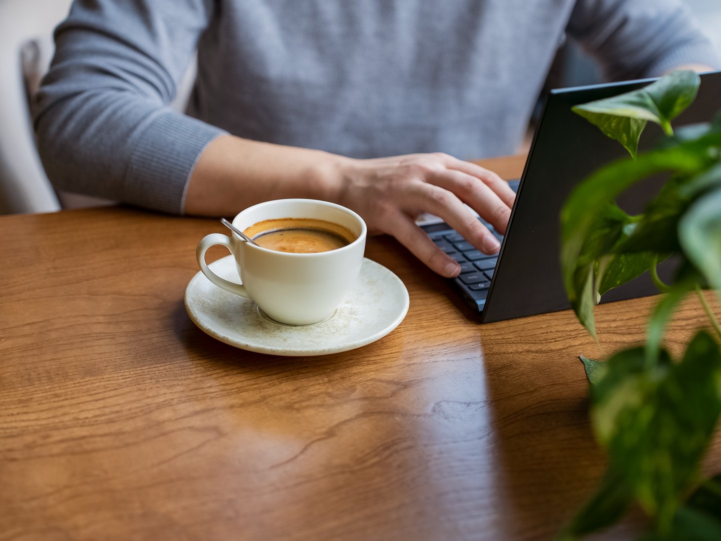 Unrecognized person in gray sweater working on laptop while sipping coffee in warm and inviting cafe. Smart freelancer sitting in restaurant with hot drink and surfing the internet.