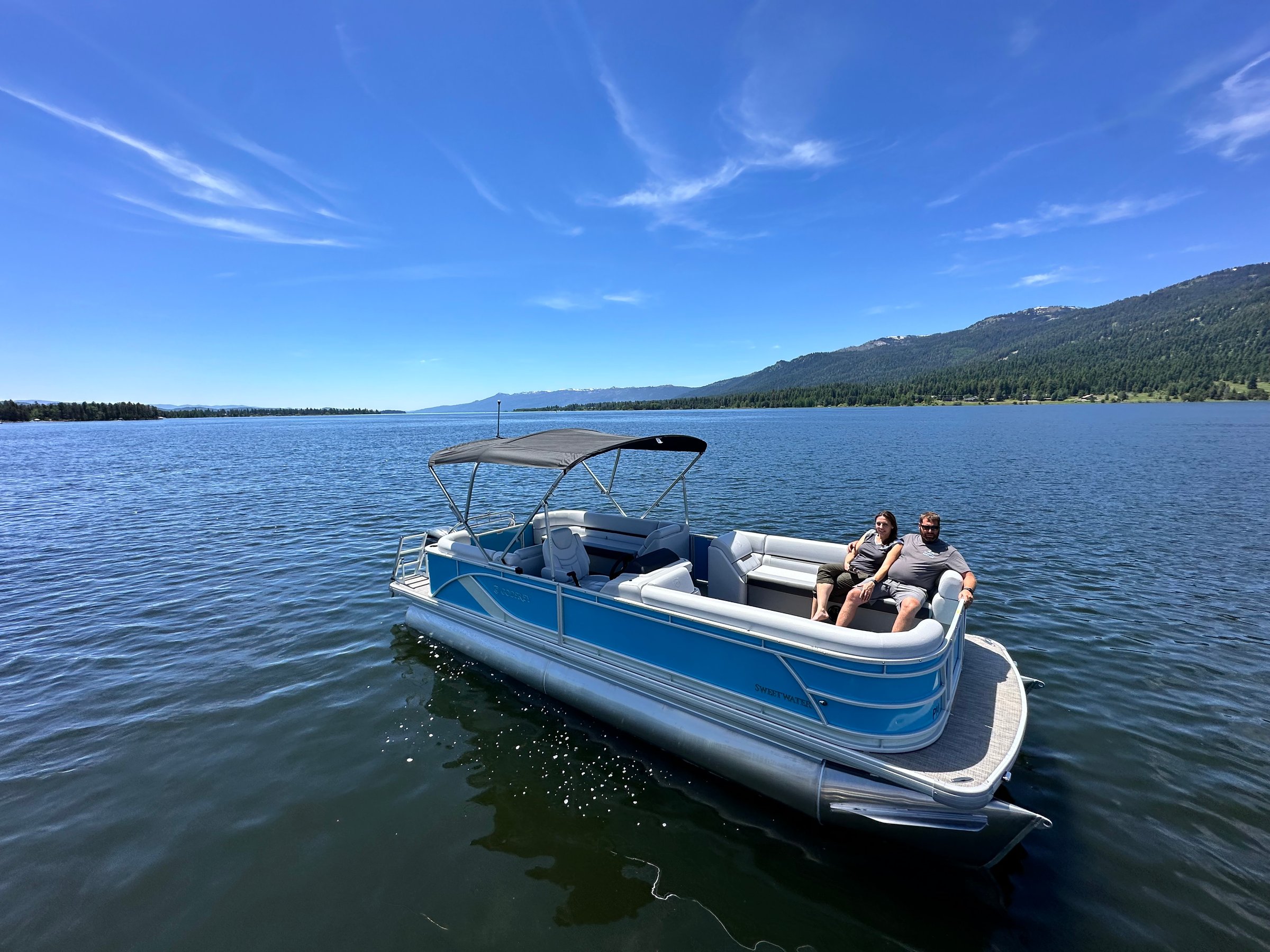A couple relaxing on a blue pontoon boat in a lake with clear skies and distant mountains.