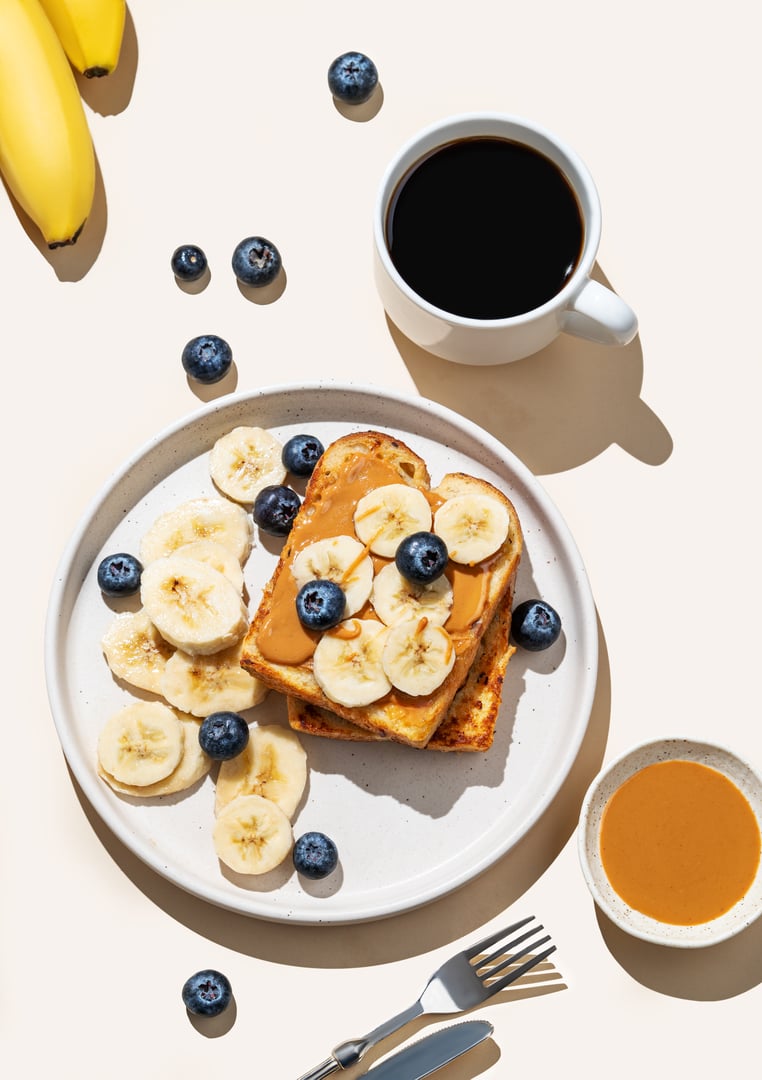 Flat lay of peanut butter toast with banana and blueberry on a plate on a beige background with a cup of espresso coffee, fruits and shadow. Concept of sweet dietary and healthy snacks for  breakfast.