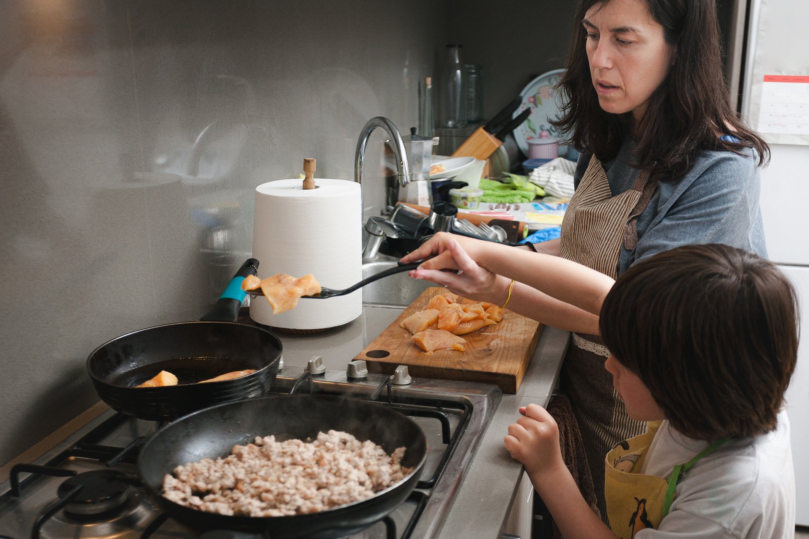 Mother and son cooking together in a modern home kitchen. The mother places pieces of raw chicken into a frying pan while the child observes attentively. A grounded and intimate moment of culinary learning and family bonding.