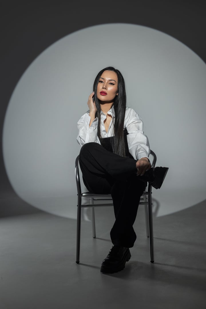 Stylish woman in black and white outfit posing on a chair in studio with dramatic lighting. Confident fashion look, long dark hair, bold makeup, minimalist and edgy atmosphere.