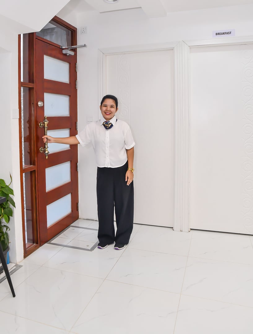 Woman in white shirt and black pants holding a door open in a hallway with white doors, tile floor, and a plant.