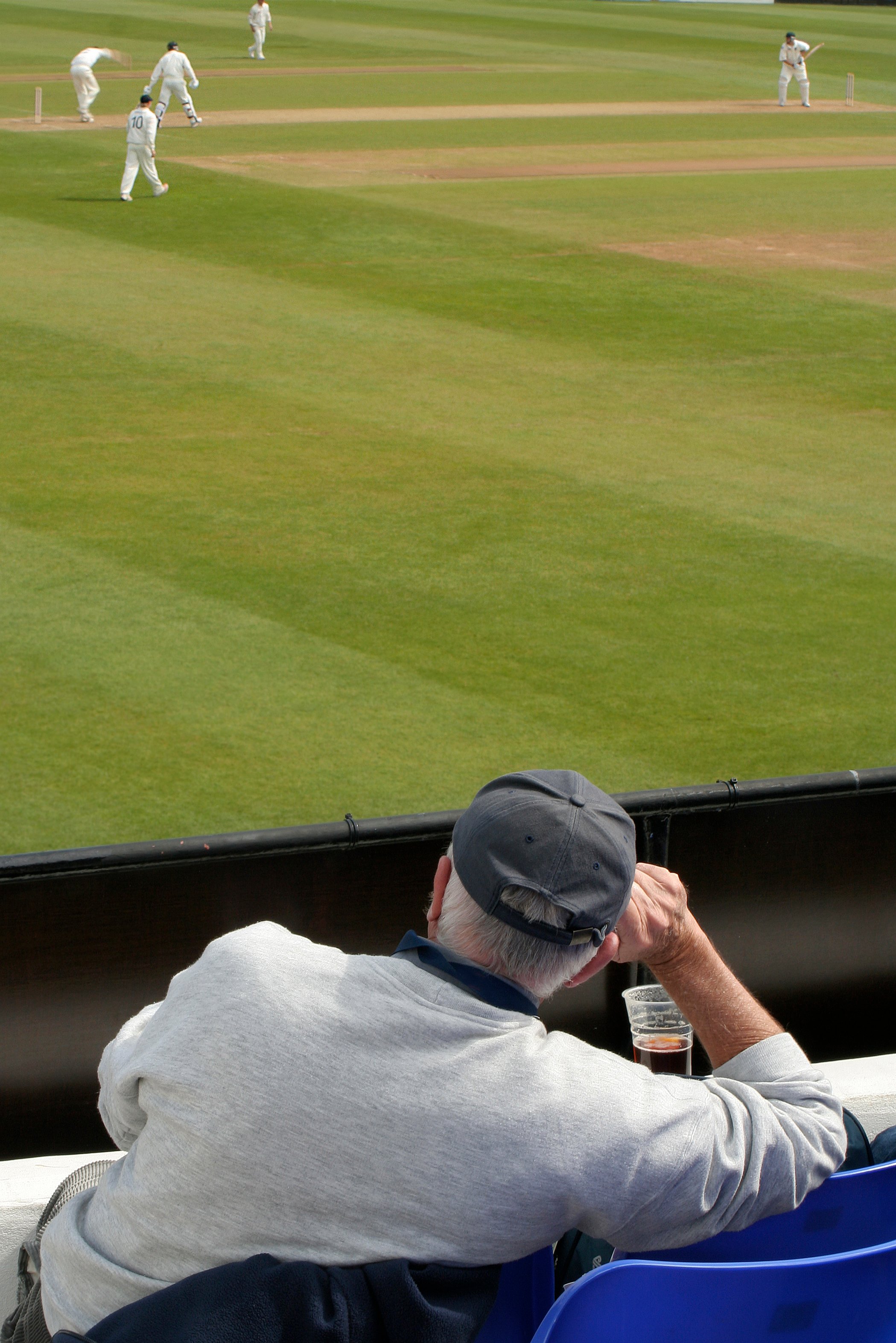 A man in a cap watches a cricket match from the stands, with players in white uniforms on the field.