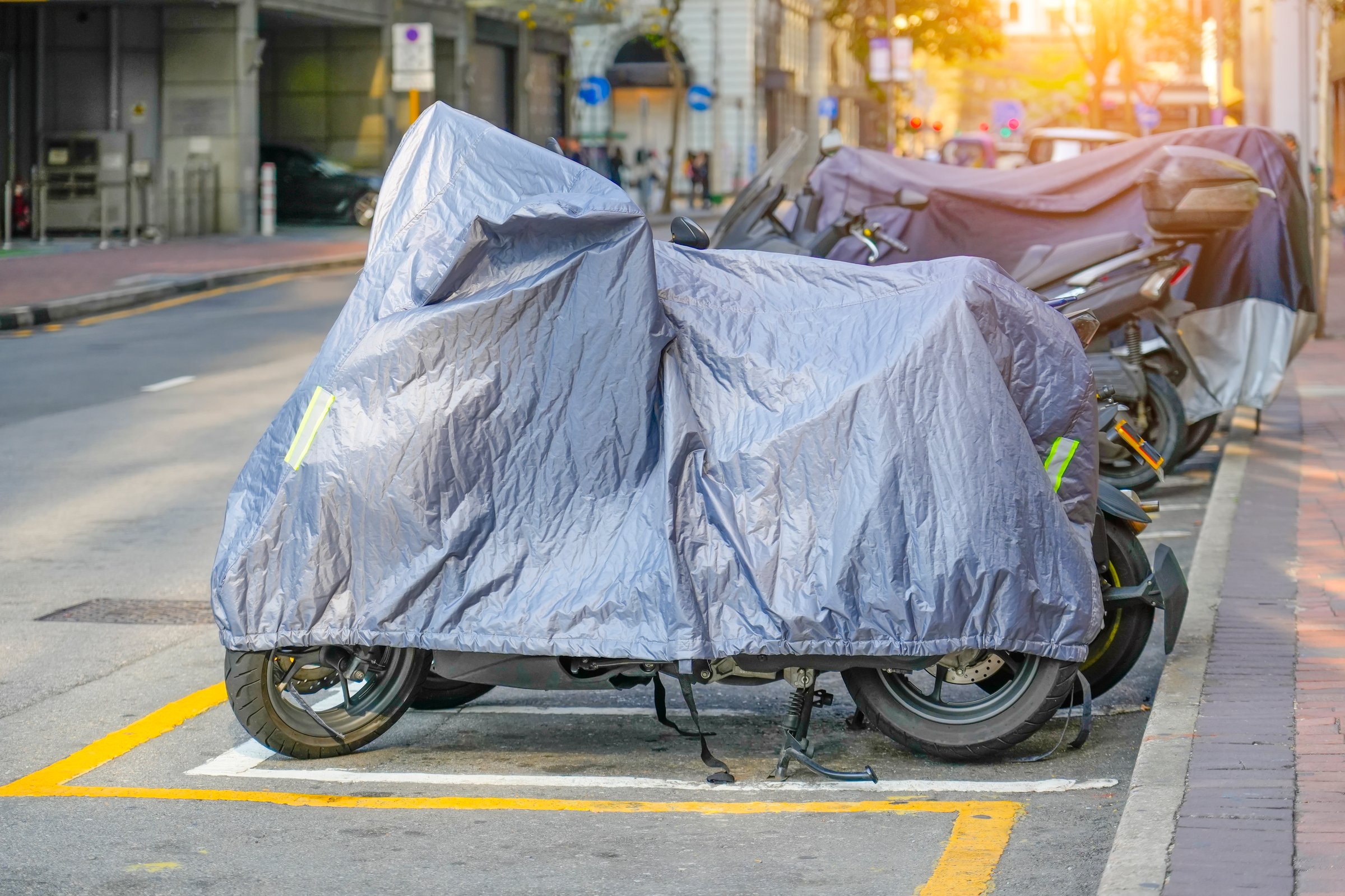 Motorcycle is covered with an gray protective jacket. A scooter with a fabric shield is parked on the sidewalk city parking. Waterproof outdoor parking while traveling.