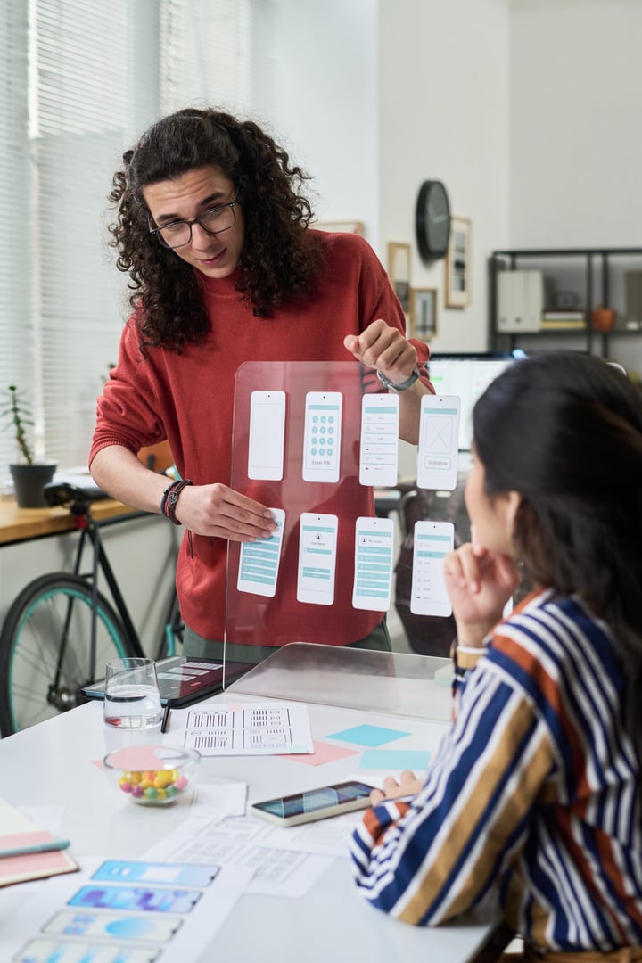 Young adult Caucasian man presenting mobile app wireframes on transparent board to young adult Latin woman sitting at desk, discussing user interface design in modern office workspace