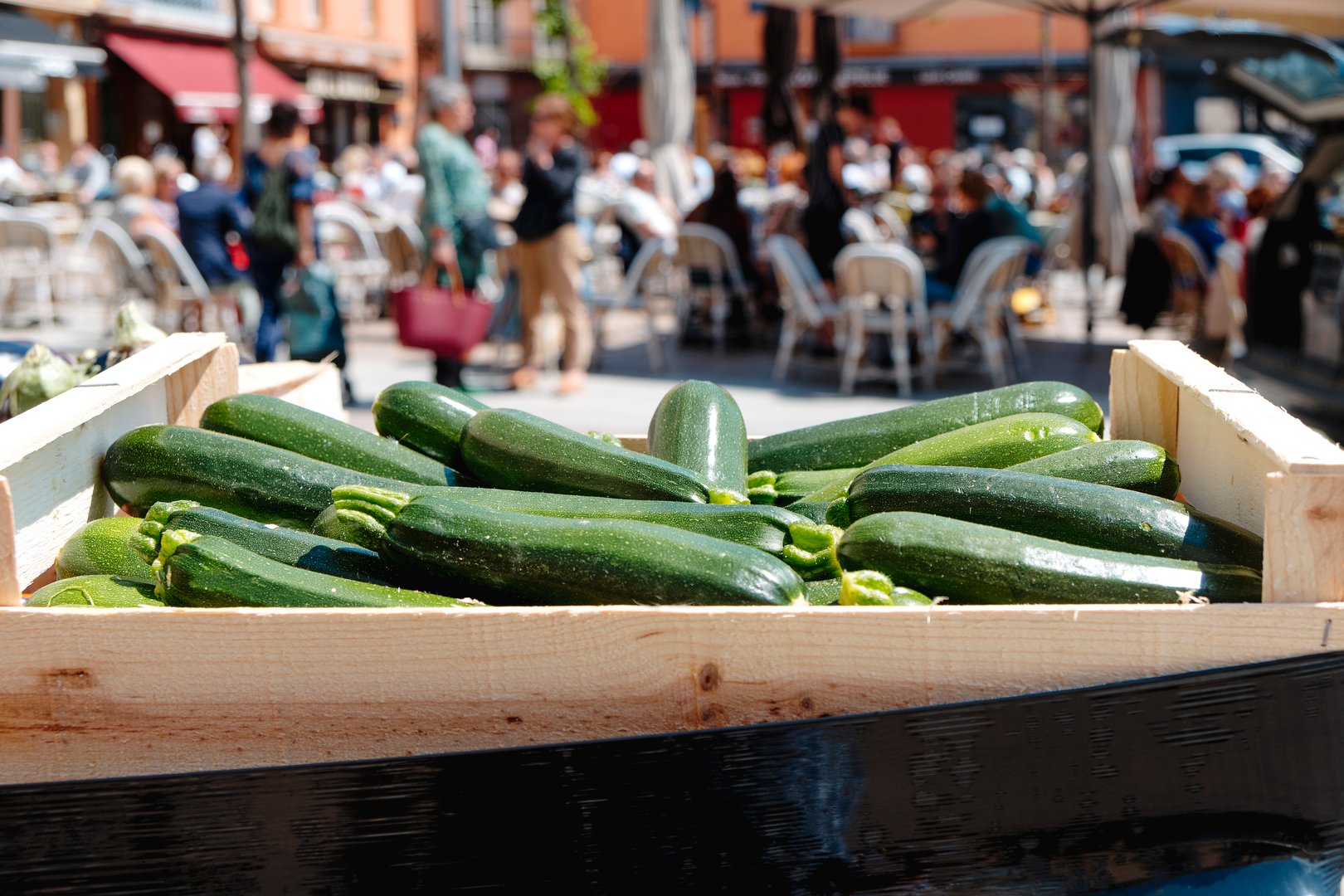 a close-up of a wooden crate with zucchinis for sale at a farmers market, with people sitting at tables of nearby sidewalk cafes in the background