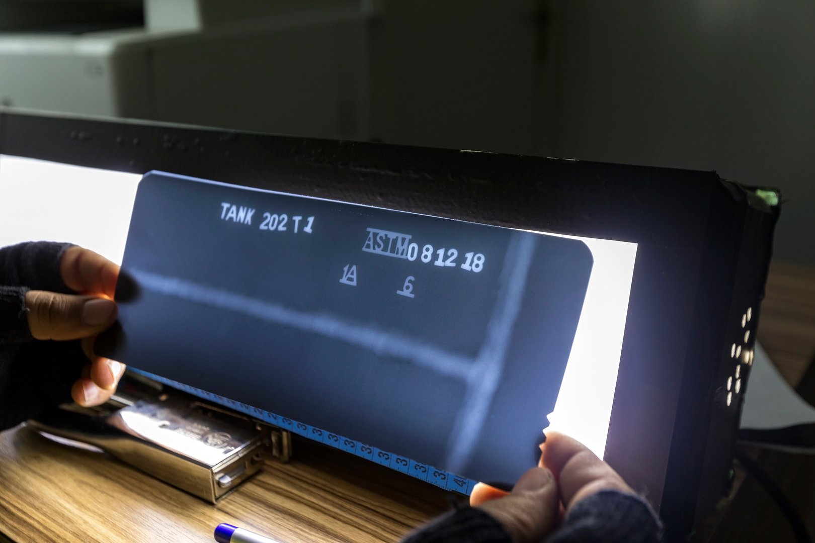 The inspector reviews radyography films of welds of the oil storage tank with light box at the office. ASTM 1A and 6 is a standard penetrameter. Tank No (202), weld no (T1) and date is a figment.